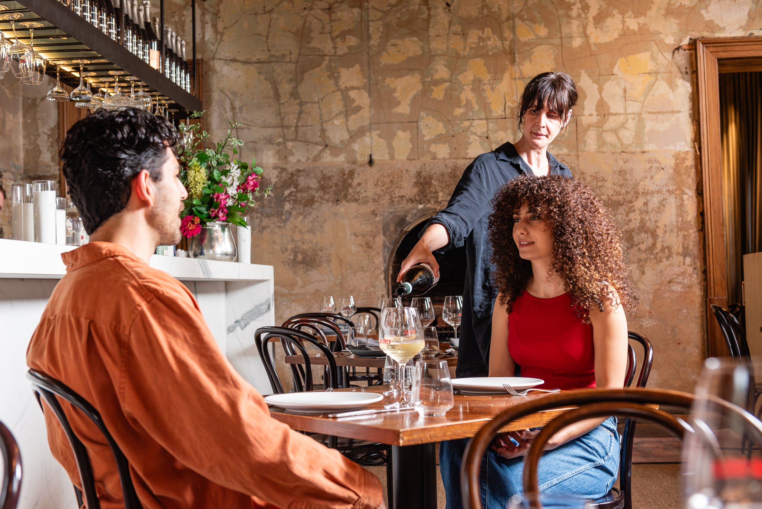 A woman in a black shirt pouring wine for a woman with curly hair and a red top at a restaurant with rustic decor, including exposed brick walls and wooden accents.