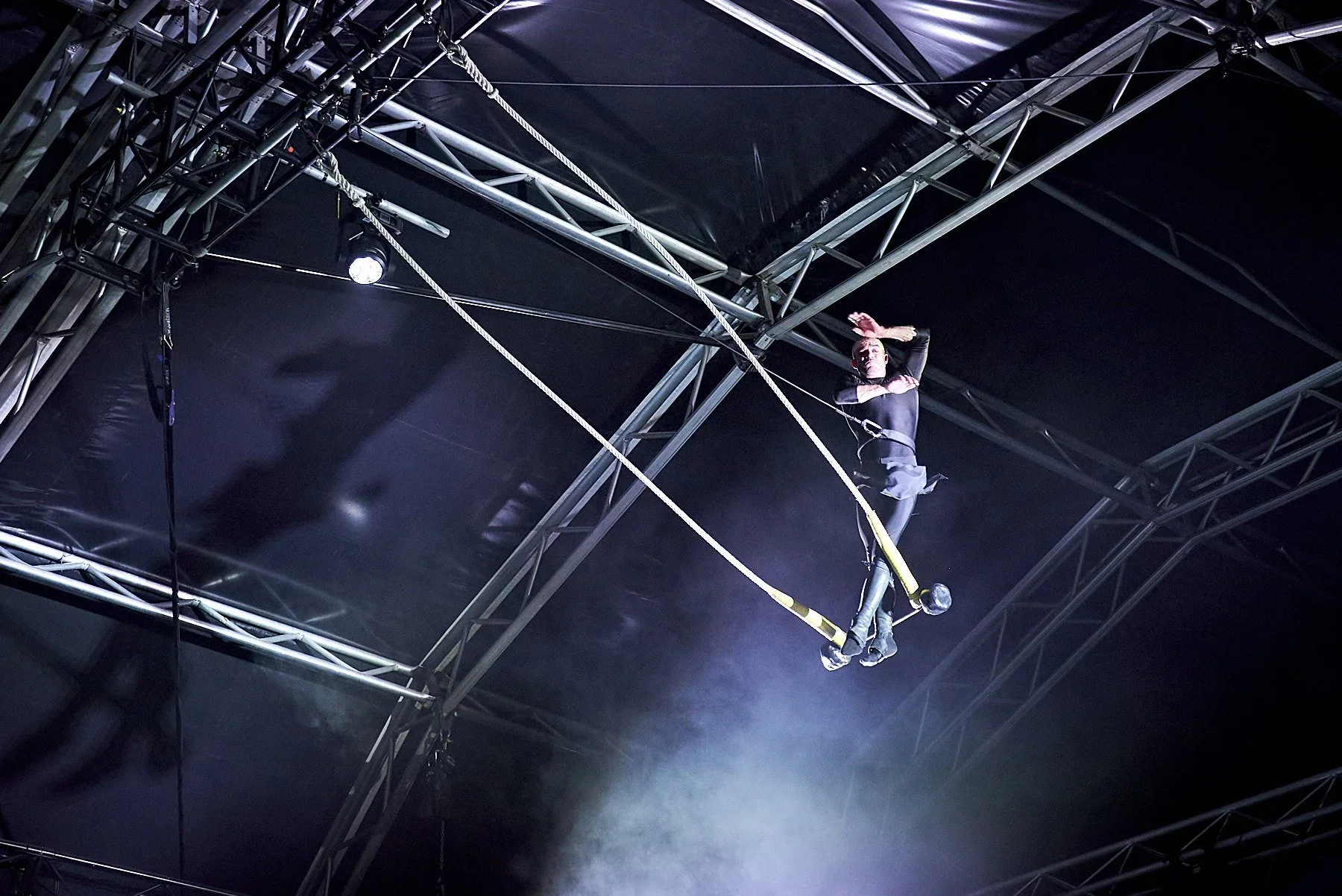 A performer on a high wire inside a circus tent, wearing black clothing, with arms raised and hands clasped above the head.