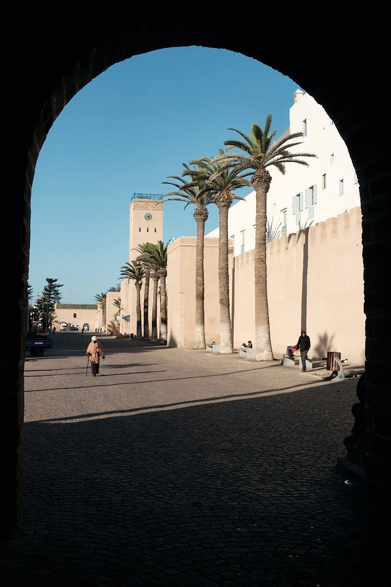 View through an archway showing a sunny courtyard with tall palm trees, a clock tower, and people sitting on benches.