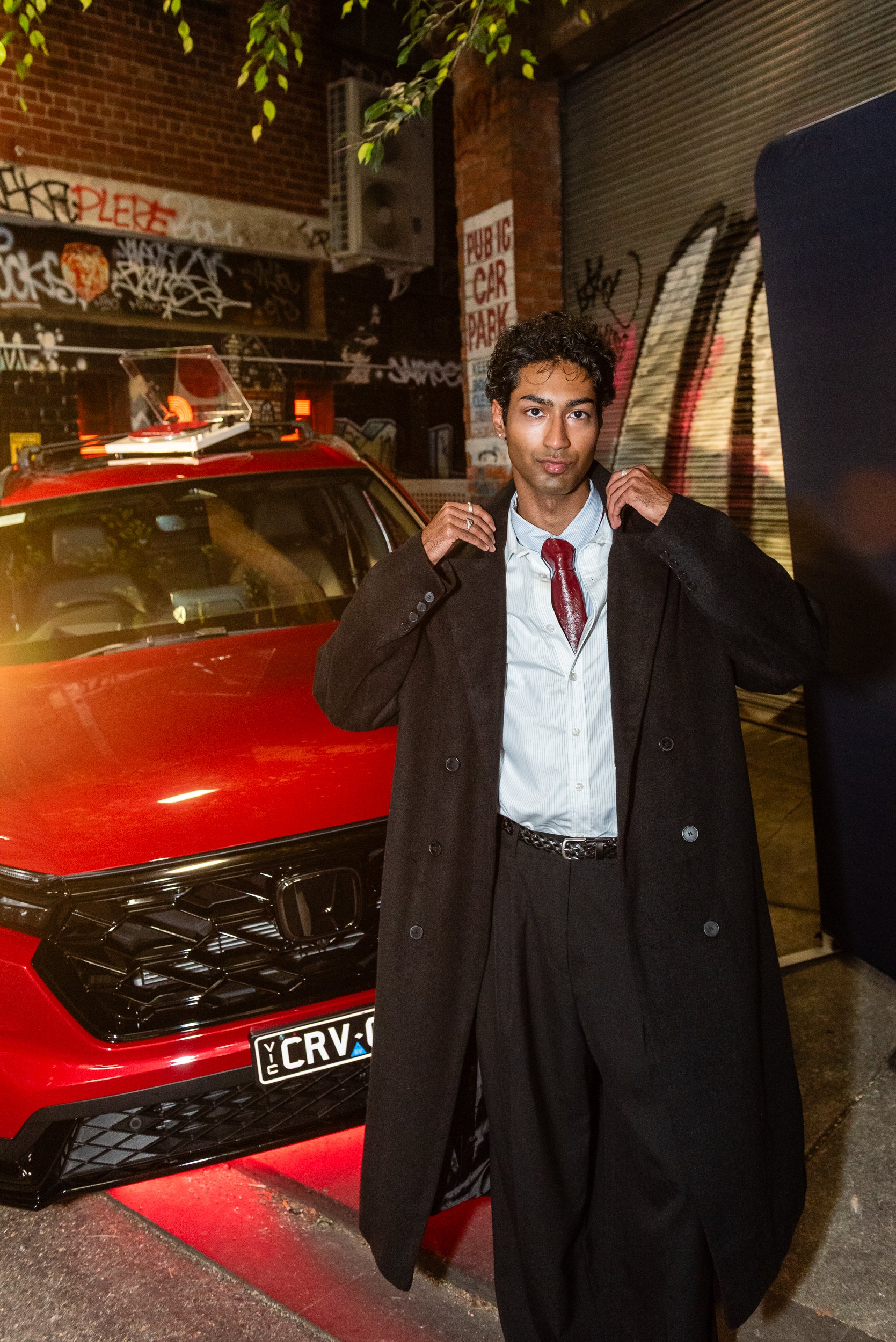 Young man in formal attire standing in front of a red Honda car with a black grille, at night in an urban alley with graffiti on brick and metal walls, and a 'Public Car Park' sign.