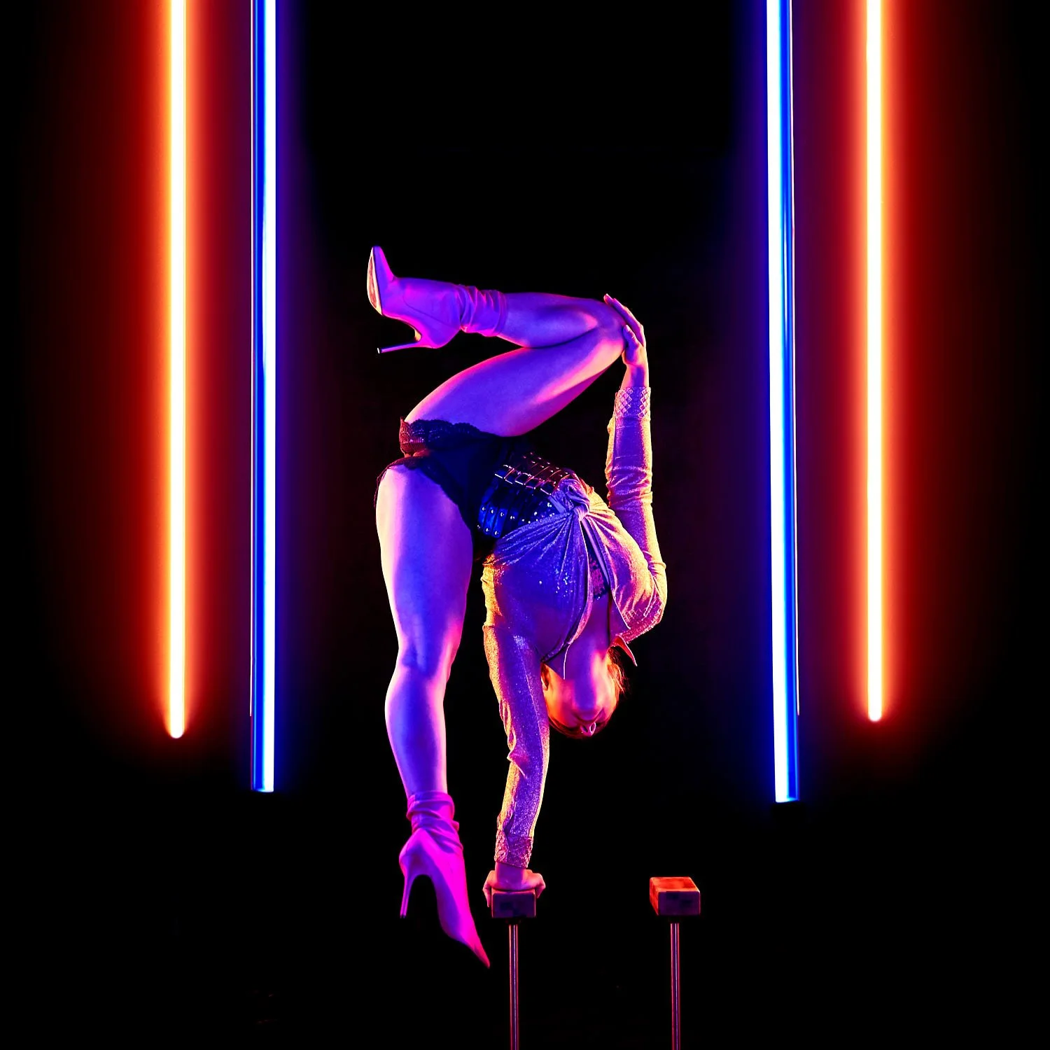 A woman performing a handstand on two elevated blocks in a neon-lit setting with colorful vertical light bars.