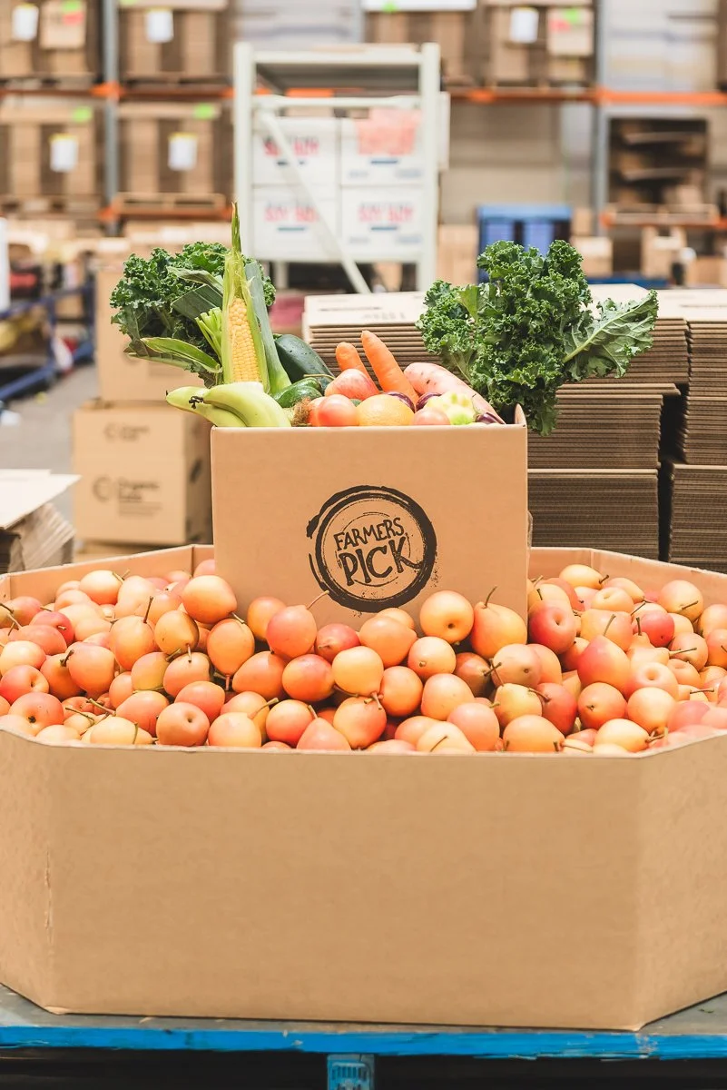 Box of small, round, pink apples with a cardboard sign reading "Farmers Pick" and assorted vegetables in the background including kale, carrots, zucchini, corn, and bananas, in a warehouse or produce section.