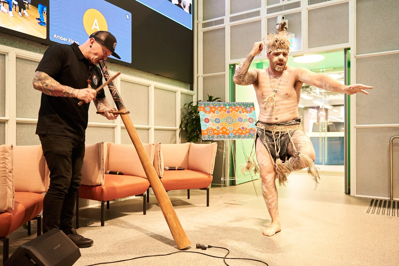 A performer with body paint, wearing tribal-like clothing, dancing during a performance, while another person plays a didgeridoo or similar instrument in an indoor setting.