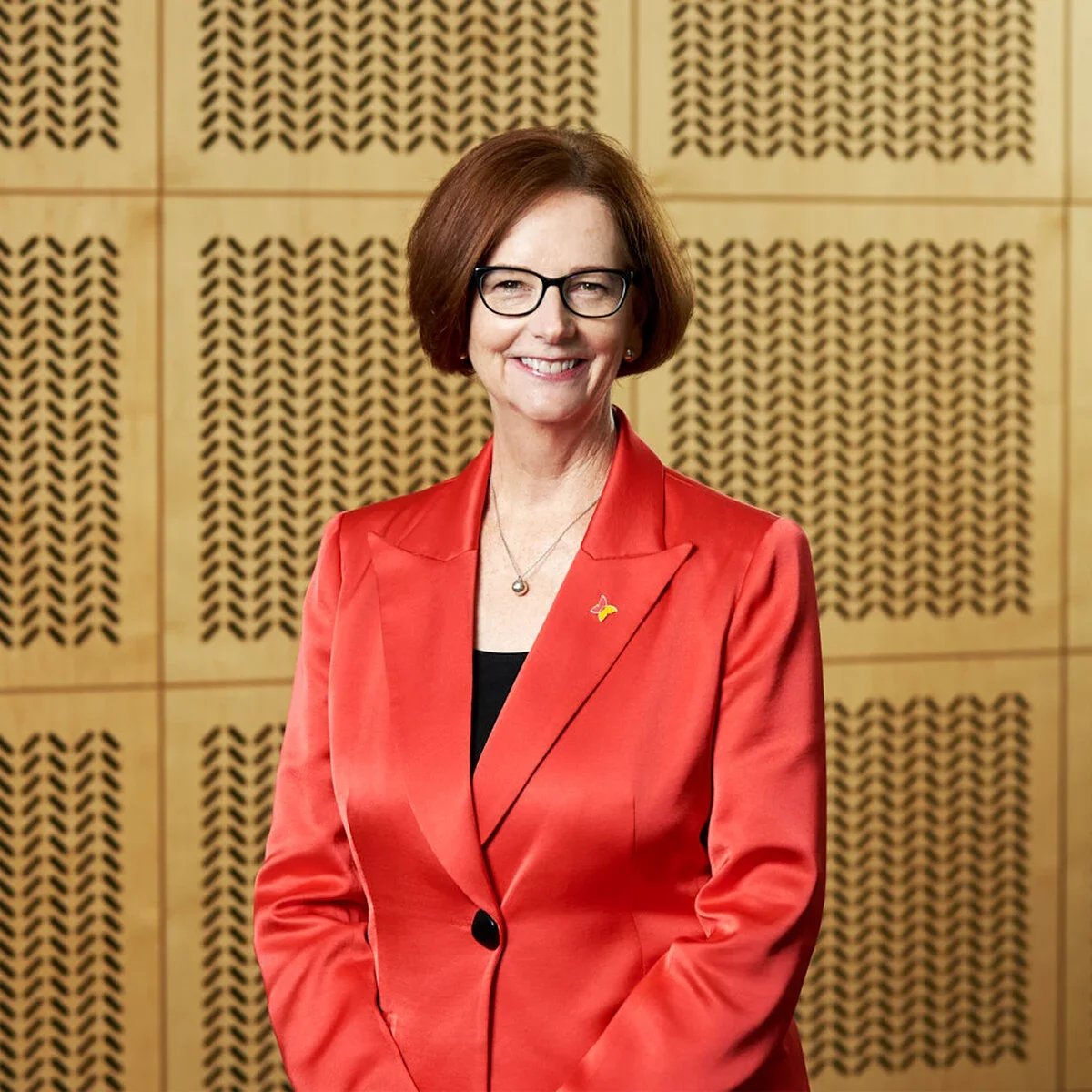 A woman with short brown hair, glasses, and a red blazer, smiling in front of a patterned wooden wall.