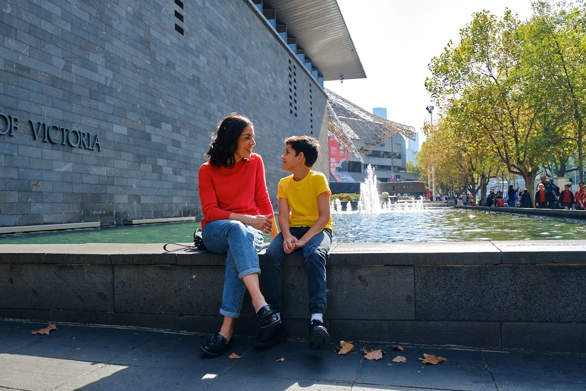 A woman and boy sitting by a fountain in an urban park, smiling at each other on a sunny day with trees and buildings in the background.