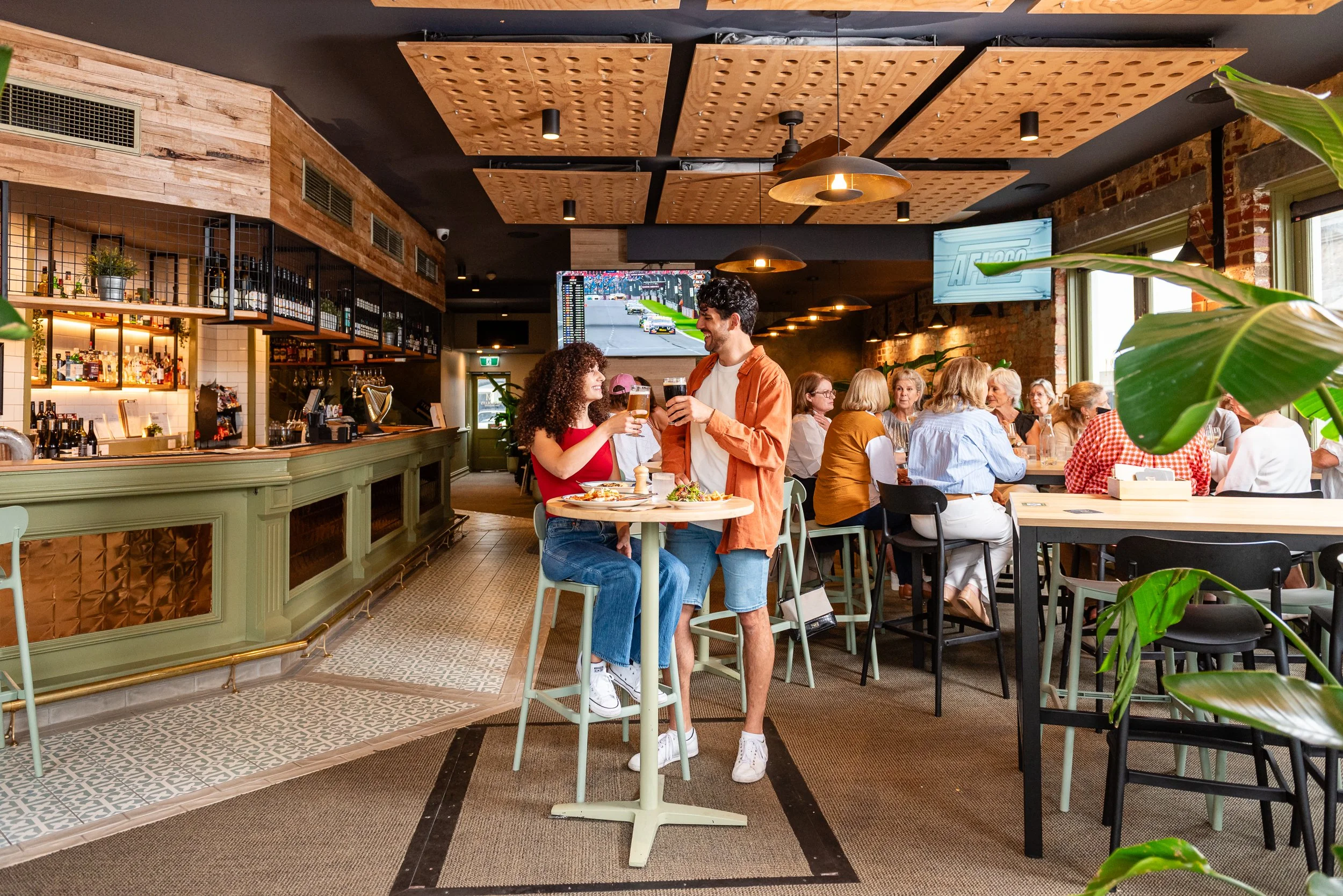 A lively bar and restaurant with a group of people enjoying drinks and conversation. Two people stand near the center, smiling and clinking glasses. The interior features wooden accents, a bar area with bottles, and a large TV screen displaying a rac