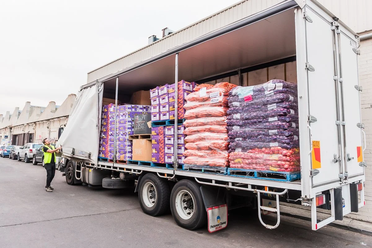 A large delivery truck with the side doors open, loaded with purple boxes, orange bags, and purple netted items, parked outside in a parking lot. A person in a yellow vest is standing next to the truck, inspecting or working.