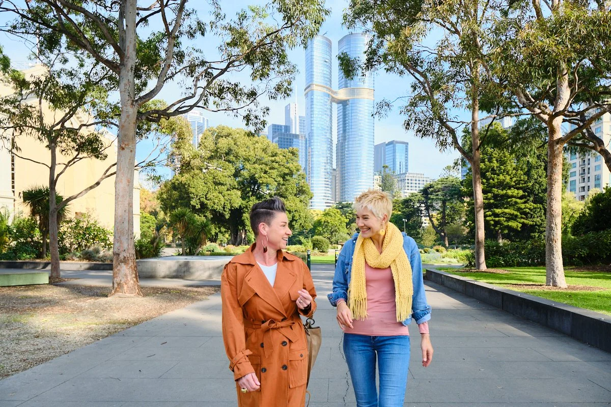 Two women walk and talk in a city park with tall modern buildings in the background. One woman wears an orange trench coat and the other a pink top with a yellow scarf.