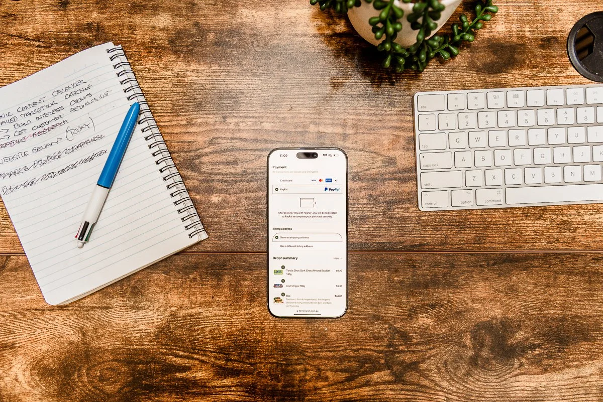 Wooden desk with a spiral notebook and blue pen on the left, a smartphone displaying an online checkout page in the center, a white computer keyboard on the right, and a plant in the top right corner.