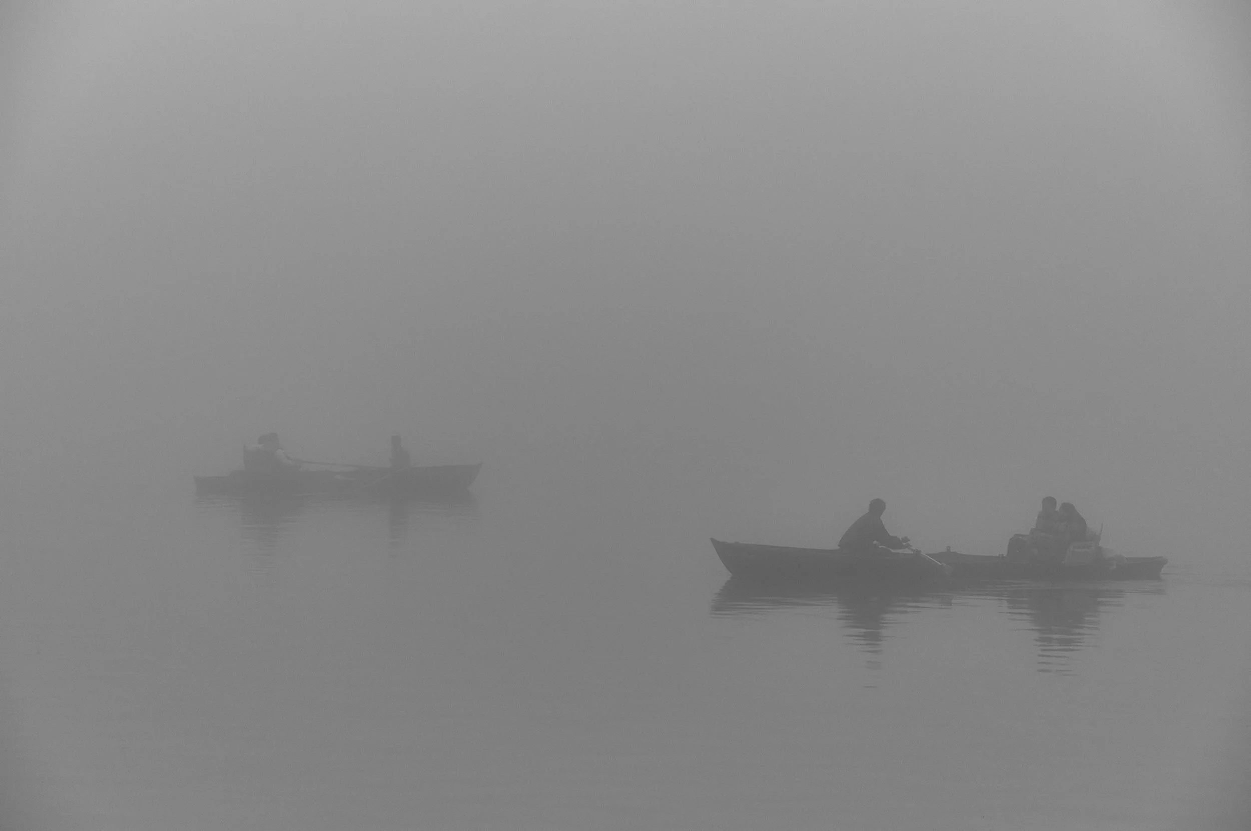 fog covered lake with two rowing boats one in the foreground one in the back ground. with people rowing slowly through the thick fog, dreamlike scene, all grey and black with reflections in the water.