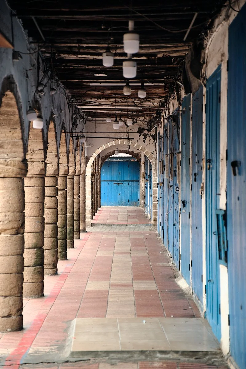 An abandoned hallway with blue wooden doors on the right and stone columns on the left, a tiled floor, and exposed ceiling with hanging lights.