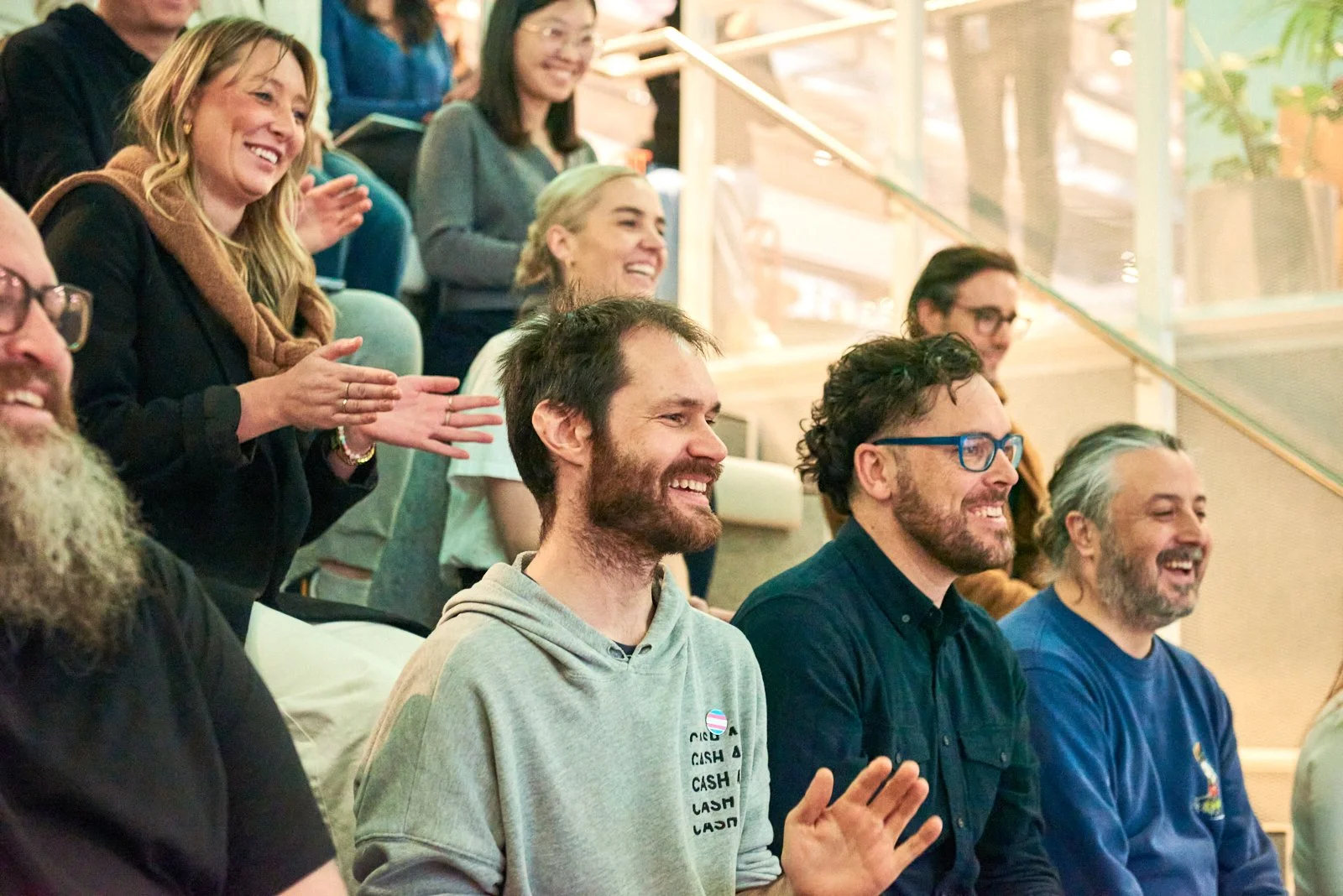 People in an audience smiling, laughing, and clapping during an event in an indoor setting.