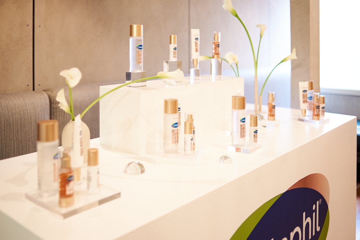 Display of skincare products arranged on a white table with white calla lily flowers in vases, featuring a large Signify logo, in natural light.