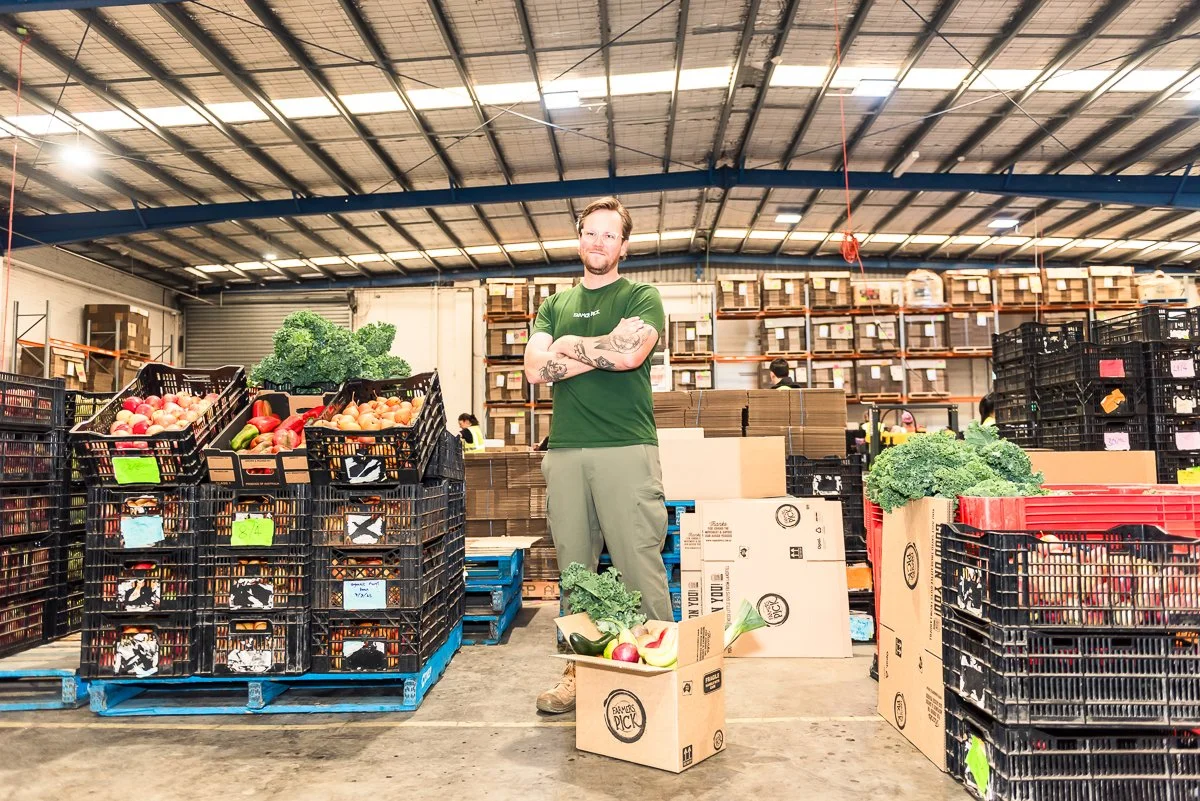 A man with tattoos wearing a green shirt and khaki pants standing with arms crossed inside a warehouse-style store, surrounded by crates of vegetables including lettuce, kale, onions, and apples.