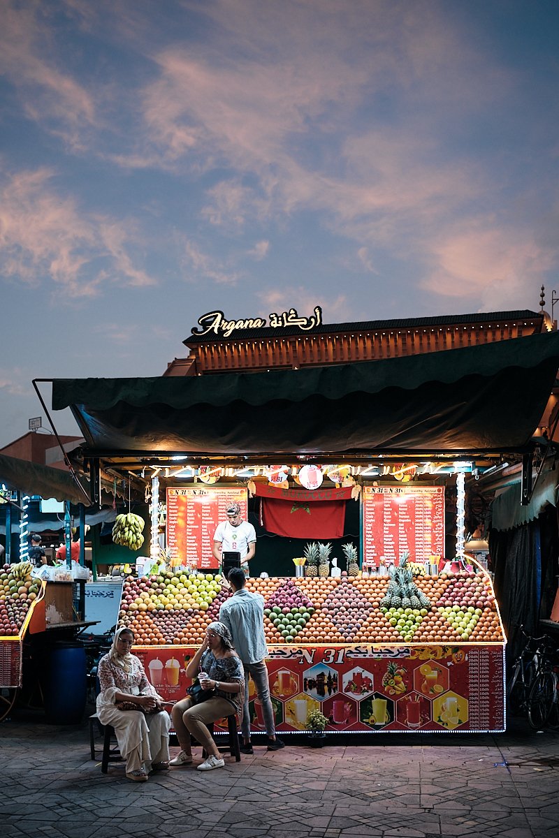 A fruit stand at an outdoor marketplace during dusk, with a sign that says "Argana" in both English and Arabic, colorful fruits displayed, and a man and two women in front of the stand.