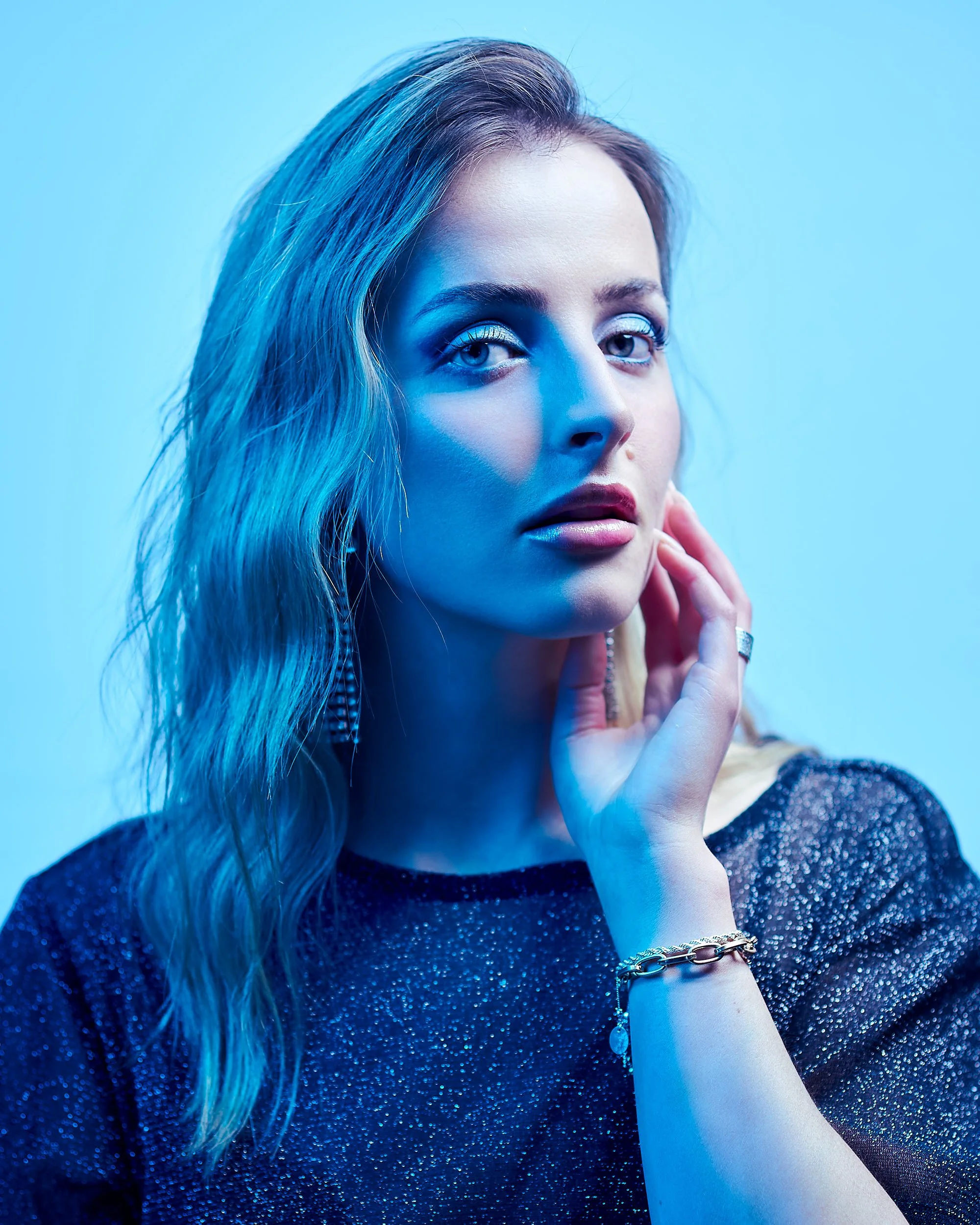 A woman with long wavy blonde hair and blue makeup, wearing a sparkly dark top, holding her face with her hand, and wearing jewelry, against a blue background.