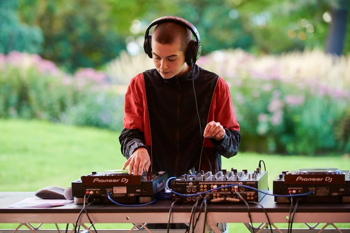 Young person with a shaved head wearing headphones, standing outdoors with greenery, operating DJ equipment on a table.