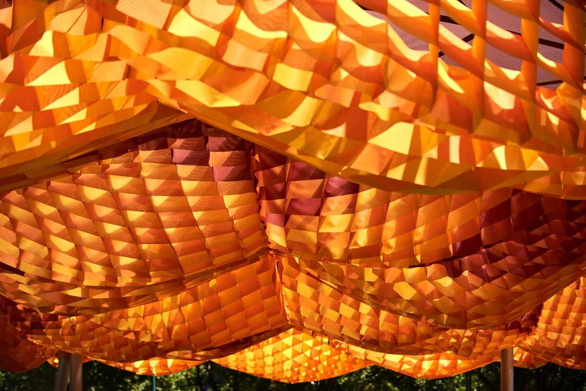 Close-up view of orange and yellow paper lanterns with geometric patterns hanging outdoors, illuminated by sunlight.