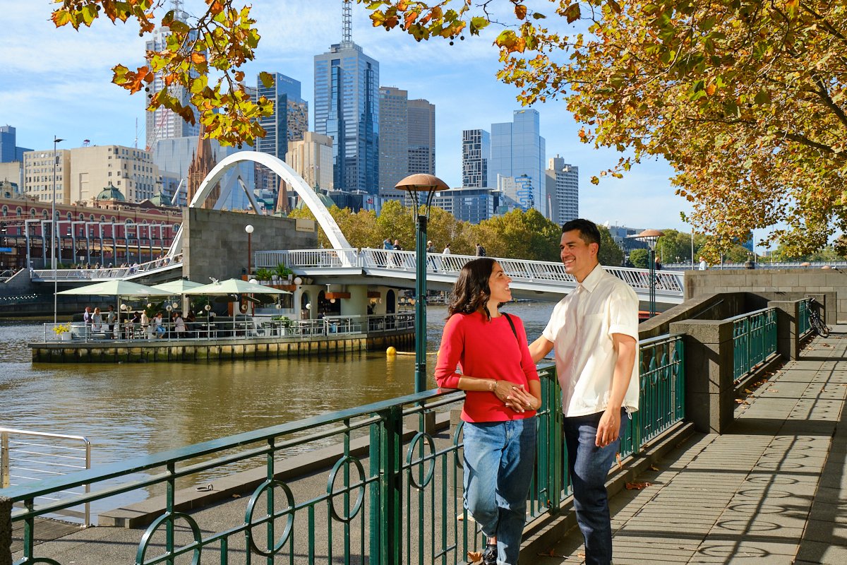 A man and woman are talking and smiling by a riverside promenade in a city with tall skyscrapers in the background. There are trees with autumn leaves overhead, a bridge across the river, and a boat docked at the riverside.