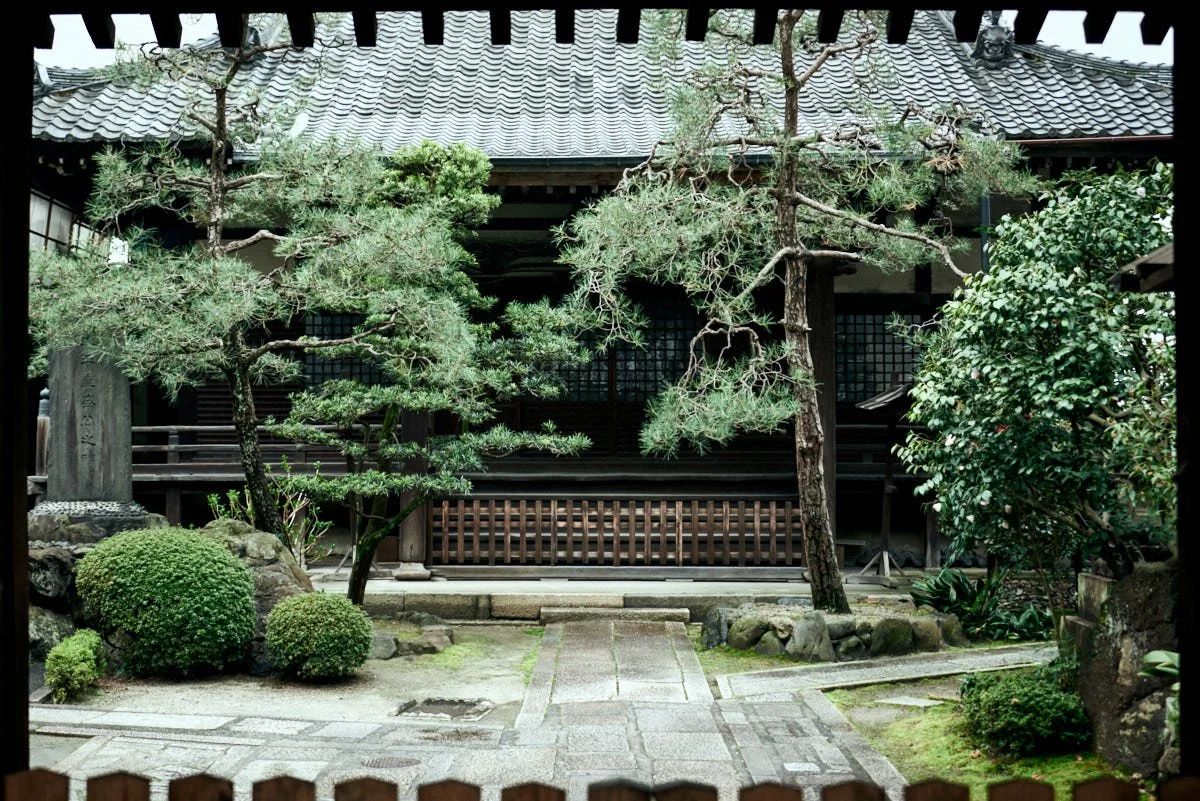 Traditional Japanese garden with trees, shrubs, rocks, and stone pathway in front of a wooden building with tiled roof.