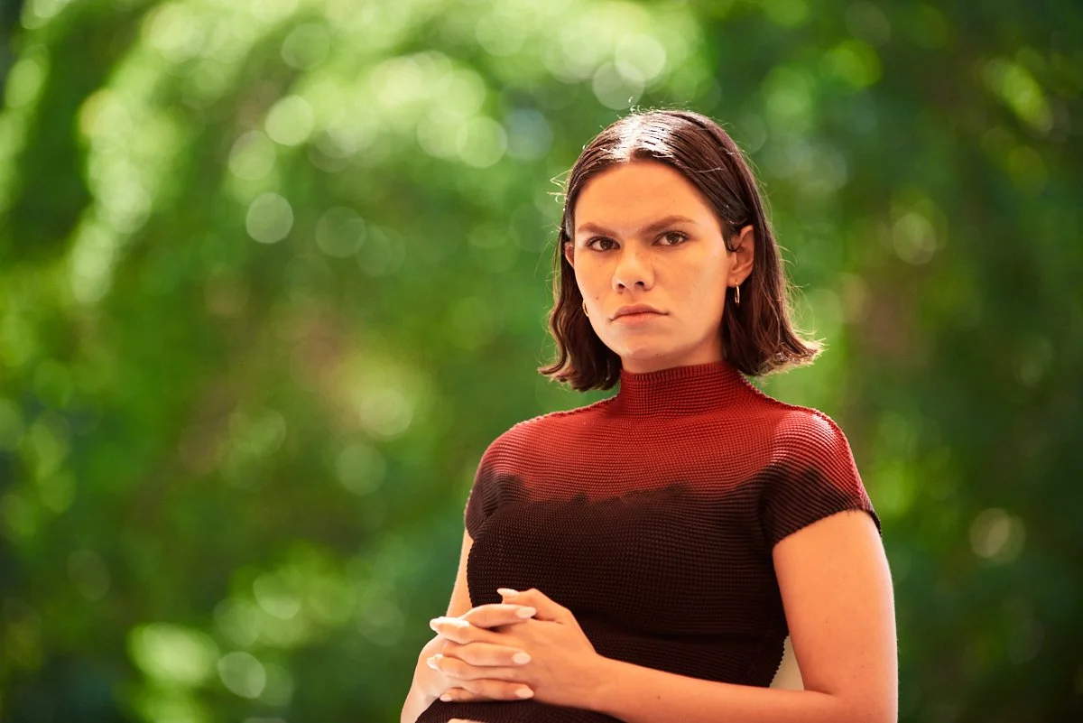 A woman with short brown hair wearing a red and black top, standing outdoors with a blurred green background.