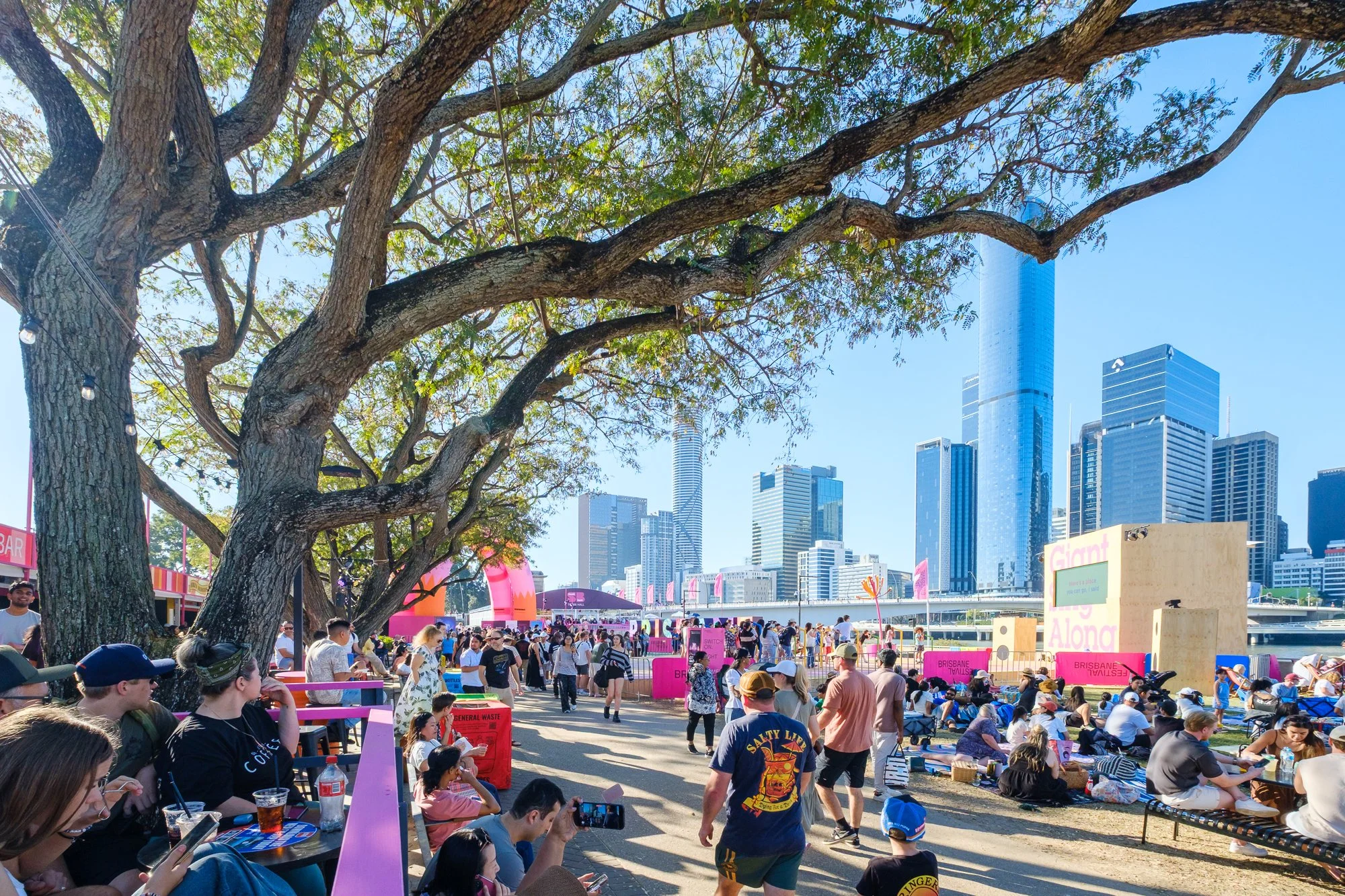 Crowd gathered at an outdoor event with pink decorations and booths, near a skyline with tall modern skyscrapers, trees, and blue sky.