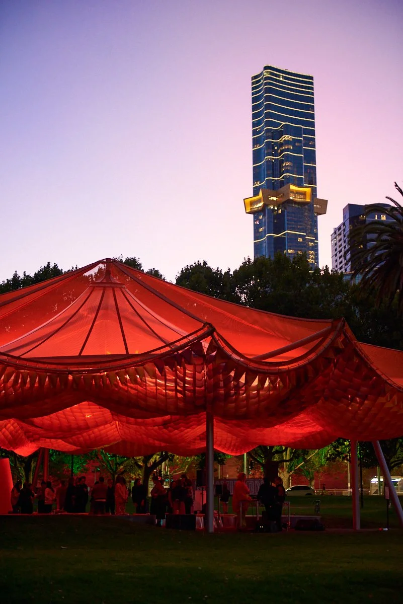 A large red tent with people gathered underneath and a tall modern glass skyscraper with blue lights in the background, during twilight.