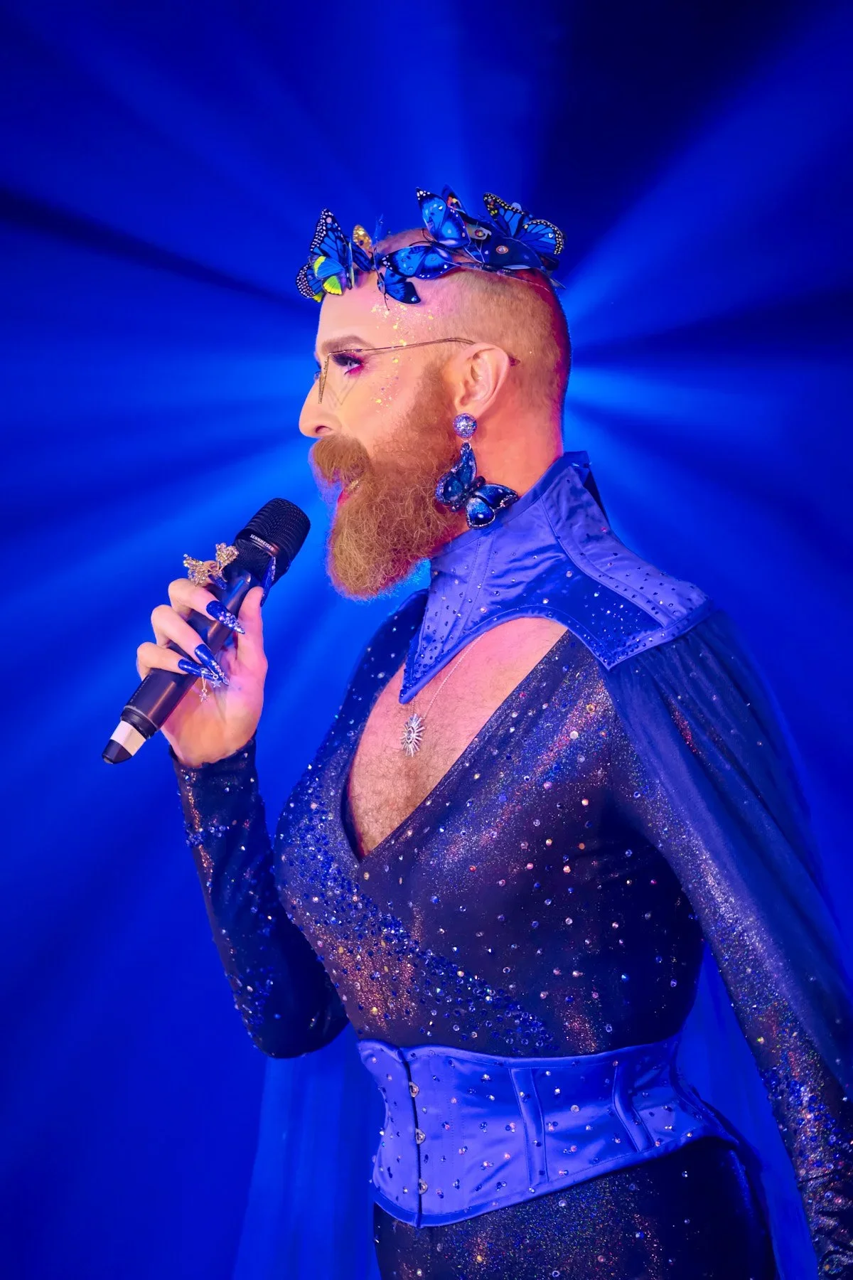 A person with a red beard and short, shaved hair performing with a microphone. They are dressed in a shiny, fitted, dark blue outfit adorned with sparkling embellishments, and wearing blue butterfly accessories on their head, earrings, and nails. Blu