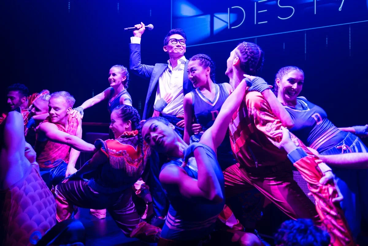 Actors on stage performing in a musical, with one man raising a microphone and others in colorful costumes.