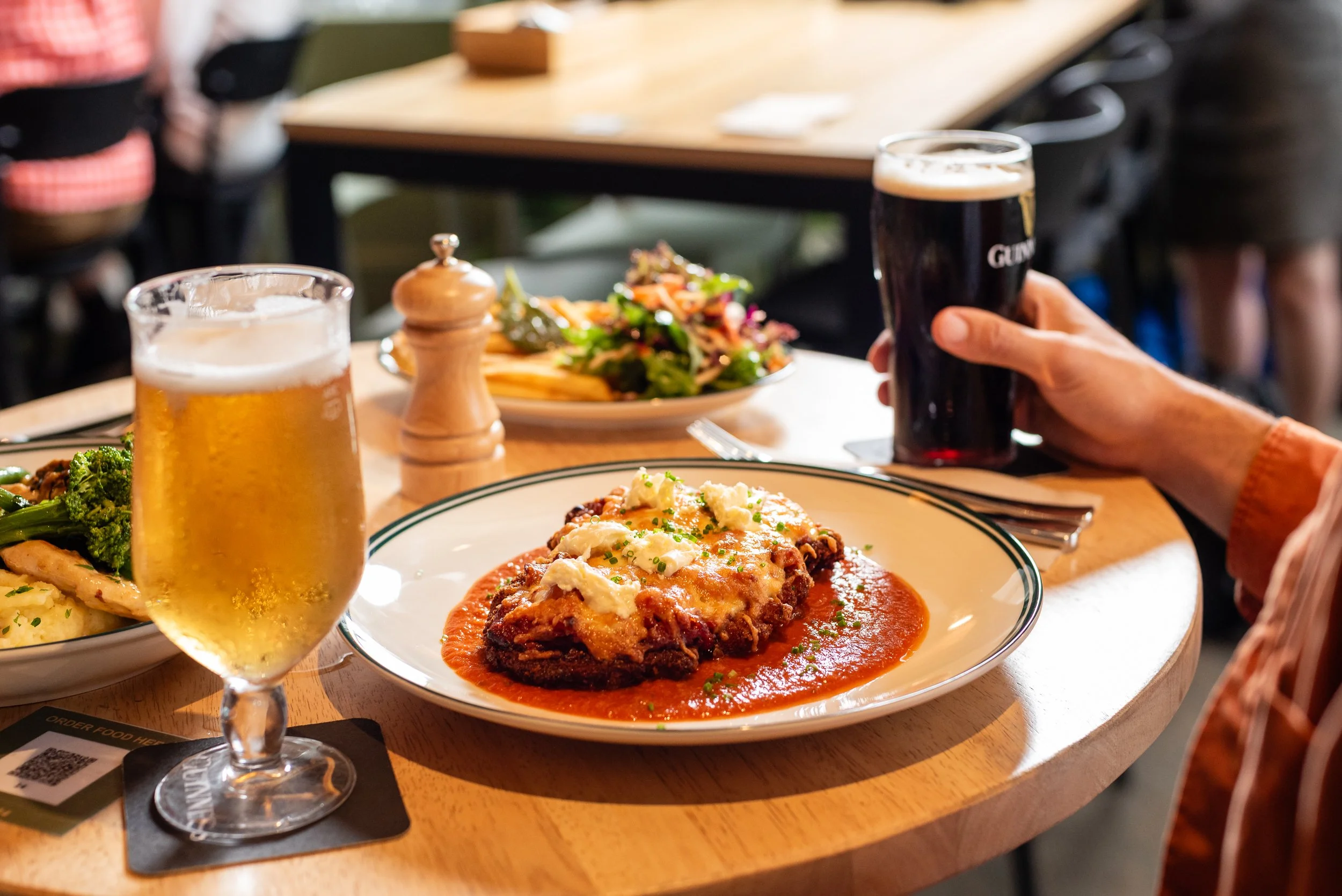 Close-up of a dinner table with a plate of Salisbury steak topped with gravy and cheese, a glass of beer, and a Plate of broccoli and mashed potatoes in the background, with a salad on a separate plate and a hand holding a glass of dark beer.