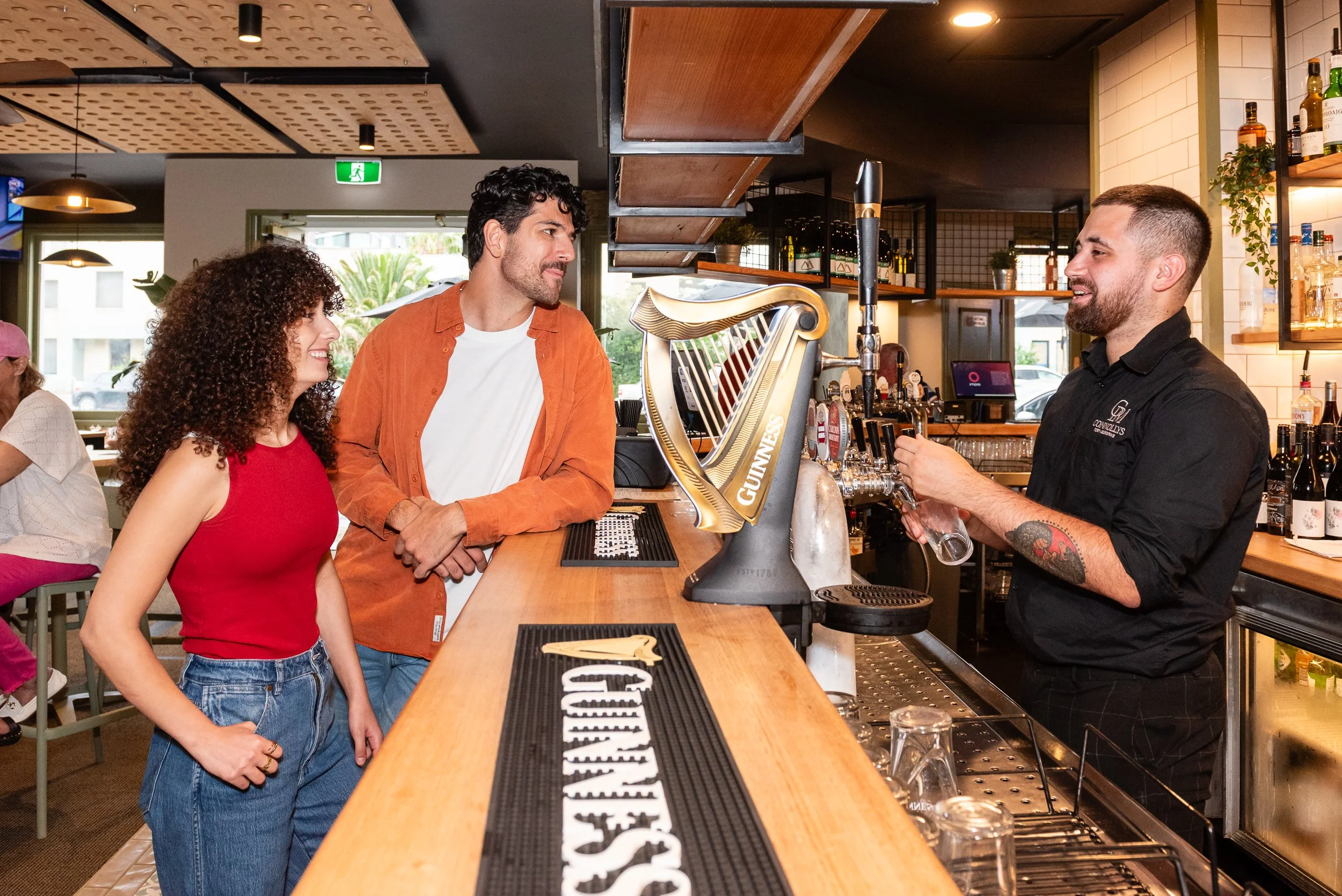 Three people at a bar talking to a bartender who is pouring a drink. The bar has a Guinness tap, and the background shows shelves with bottles and glasses.