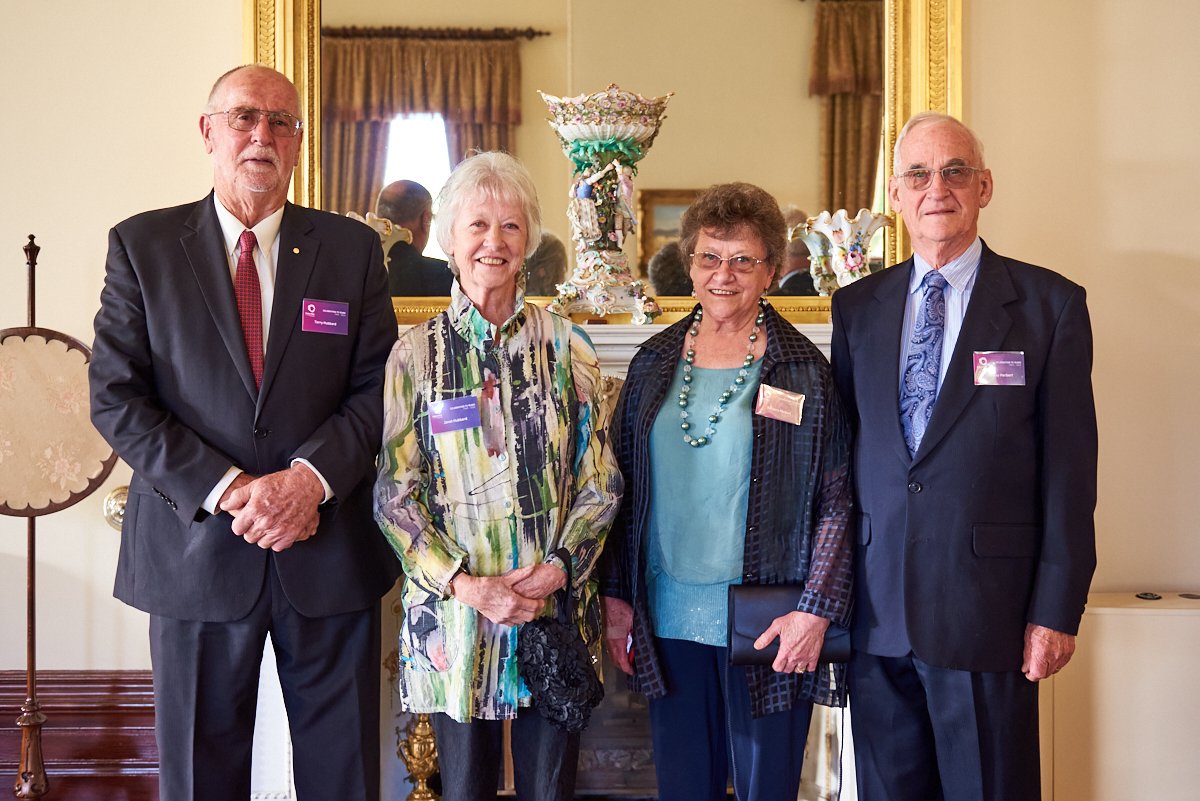 Four older adults, two men and two women, standing indoors in front of a mirror and ornate decorative porcelain vase, dressed in formal attire, smiling for a photo.