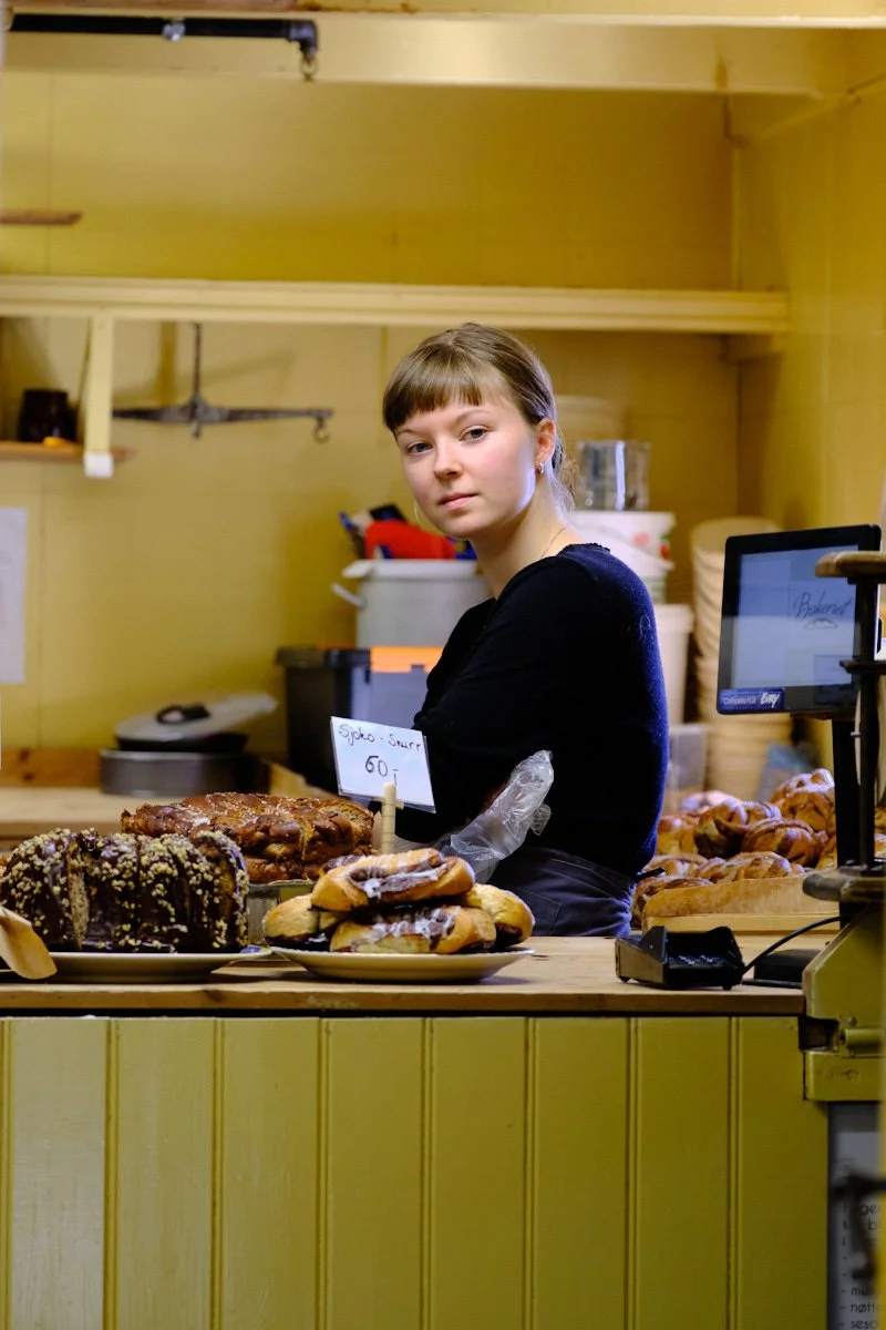 A young woman standing behind a bakery counter with various baked goods, wearing a black sweater and a plastic glove on her left hand. The bakery has a yellow wooden interior with shelves and boxes in the background.