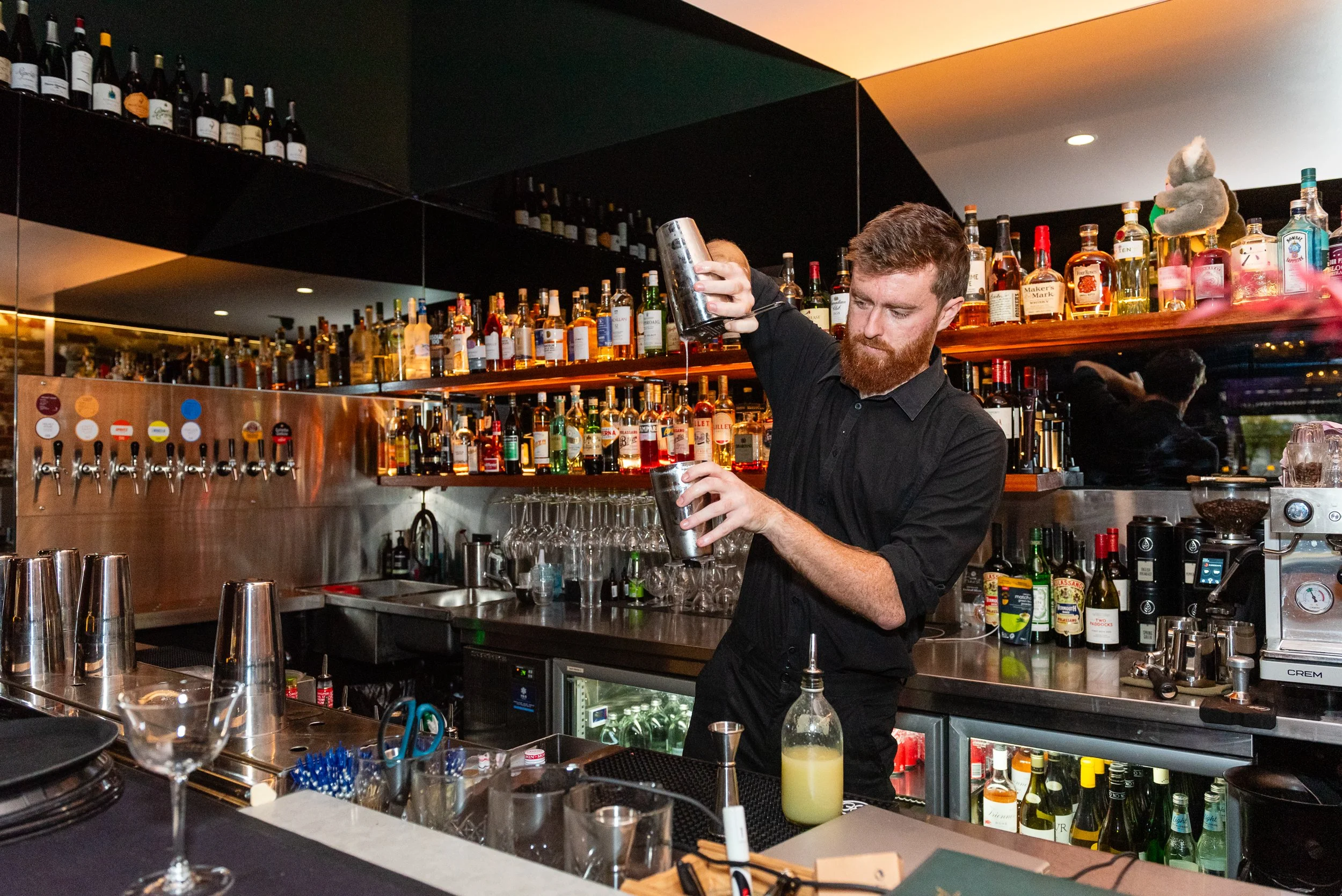 A bartender with a beard pouring a drink into a shaker at a bar with bottles of alcohol lined up on shelves behind him.