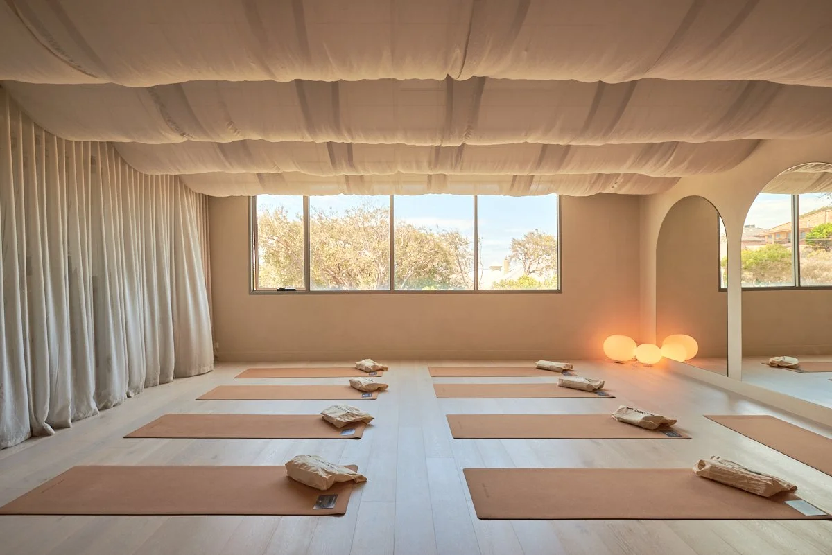 A spacious yoga studio with eight brown mats and folded towels, large windows letting in natural light, soft glowing round lamps in the corner, and a large mirror on the wall.