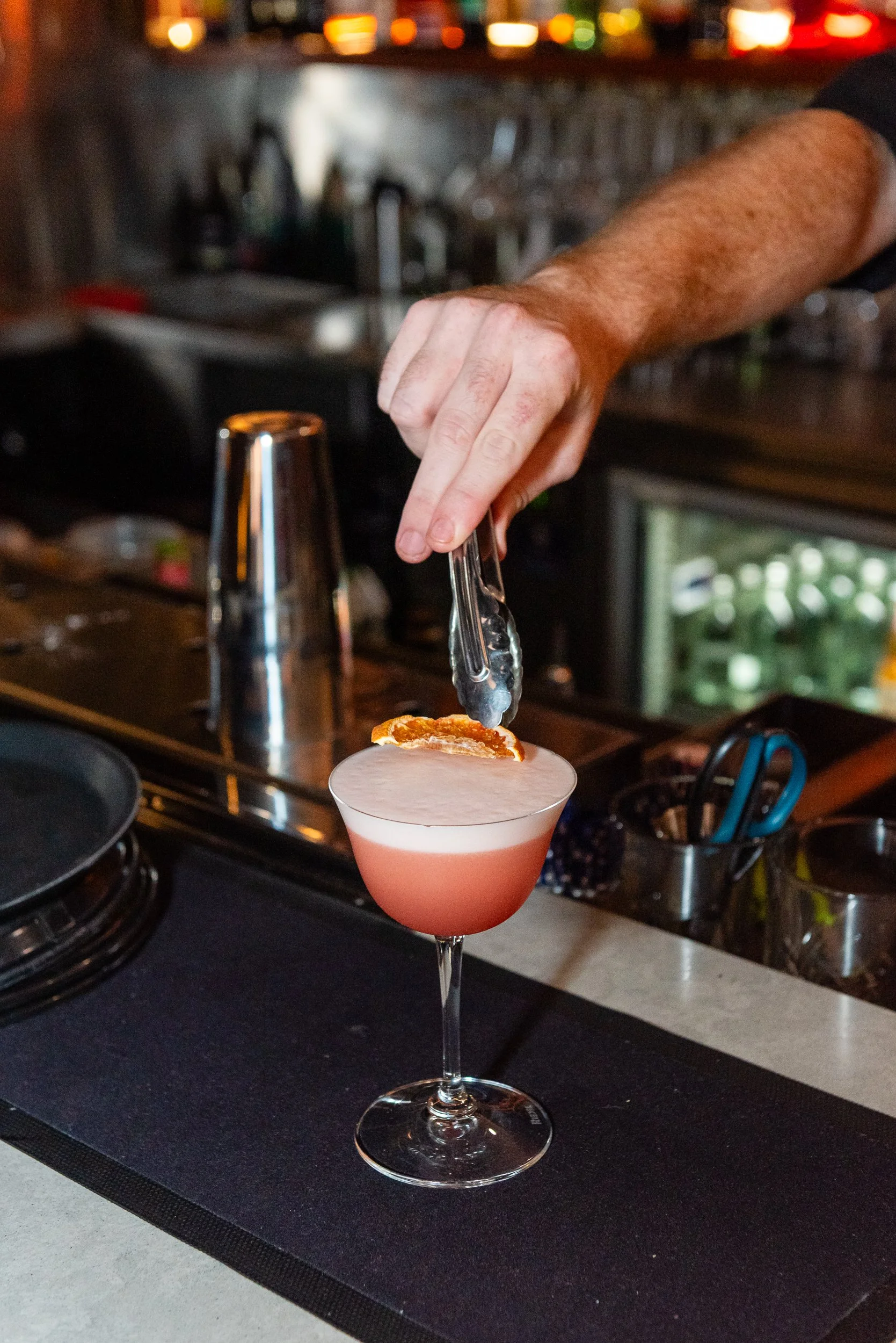 A person using tongs to place an orange slice into a pink cocktail in a coupe glass, on a bar counter with kitchen appliances in the background.