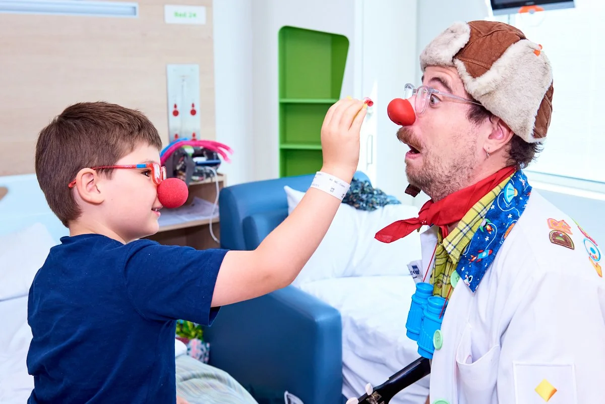 A young boy and an adult dressed as a clown, both wearing red clown noses and glasses, engaging in a playful activity in a hospital room. The boy is touching the clown's nose.