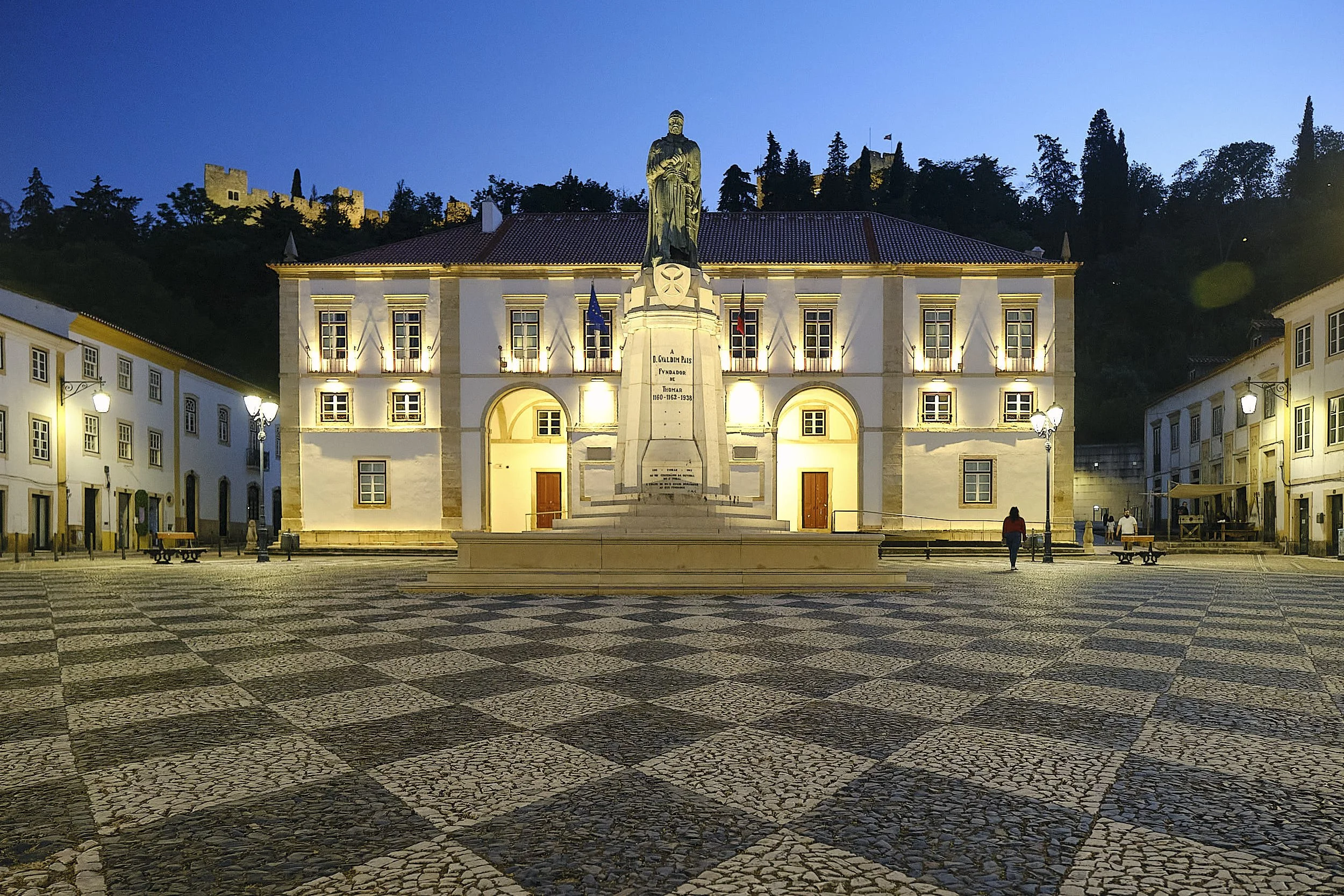 A town square at dusk with a large illuminated building featuring a statue in front. The statue stands on a pedestal and depicts a figure, possibly historical or religious. The square is paved with patterned tiles and has street lamps and a few peopl