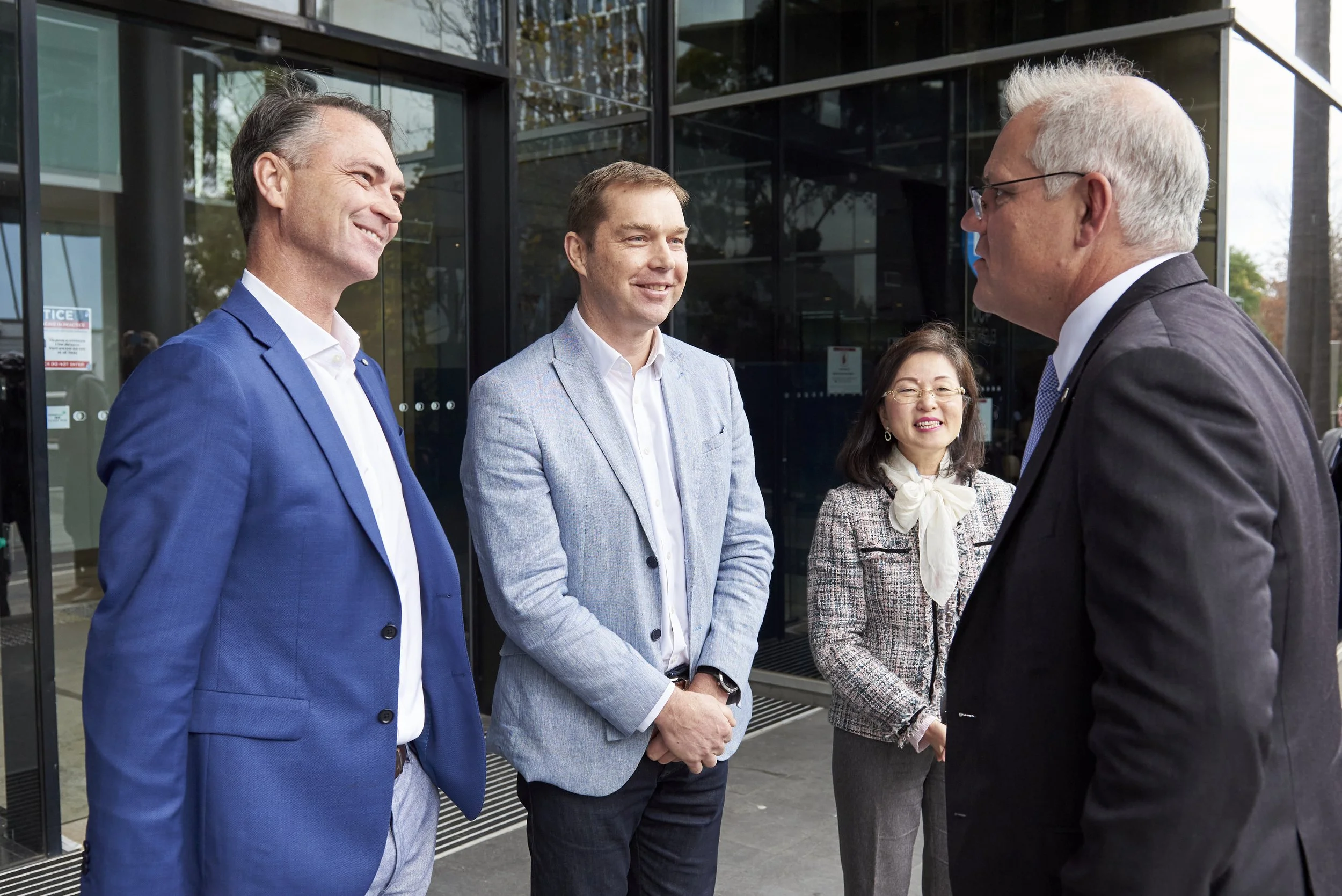 Four business professionals, three men and one woman, engaged in conversation outside a modern glass office building. The men are dressed in suits, and the woman is wearing a tweed jacket with a bow tie.
