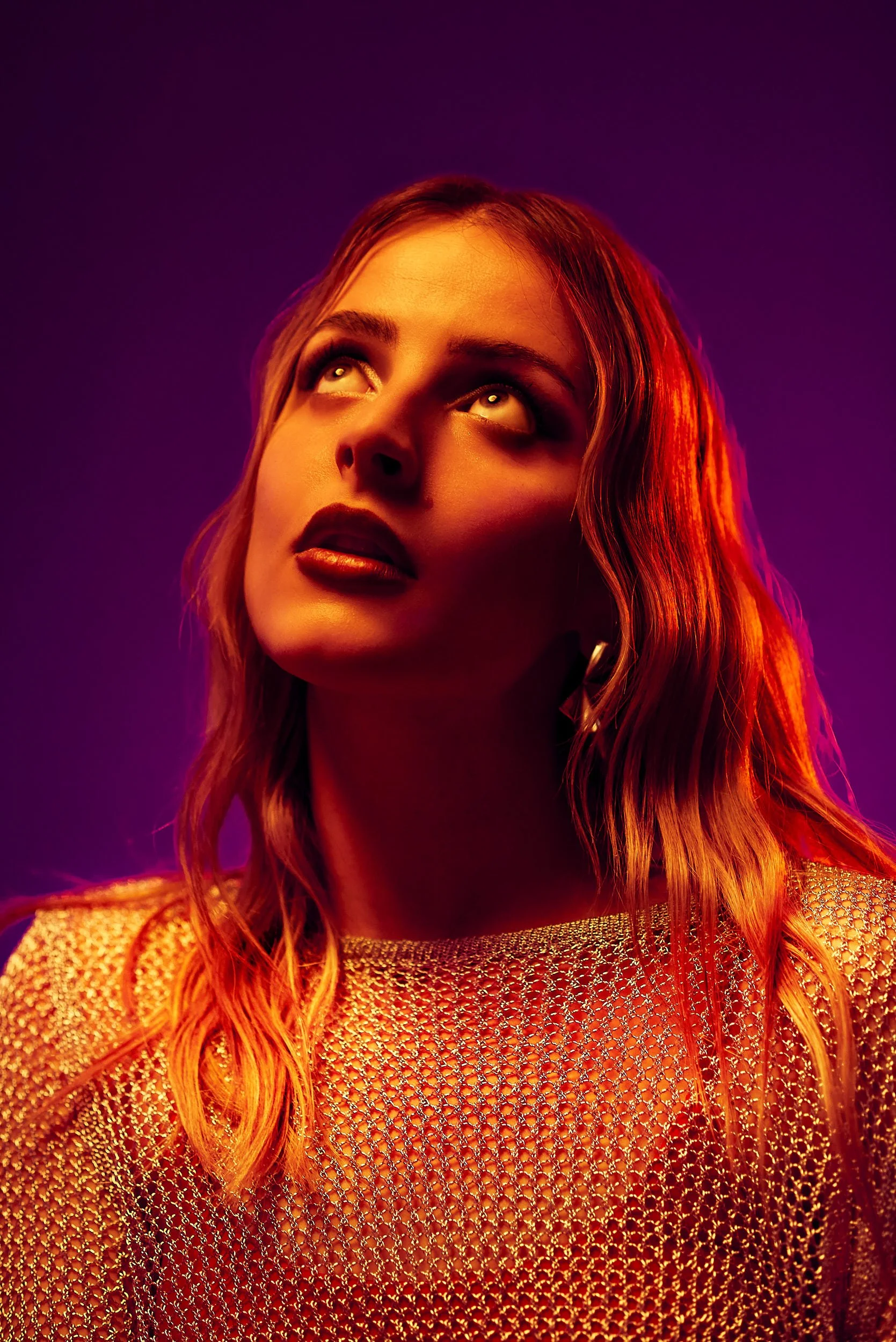 A woman with wavy hair looking upward, illuminated with warm orange and purple lighting, wearing a sparkling, netted top.