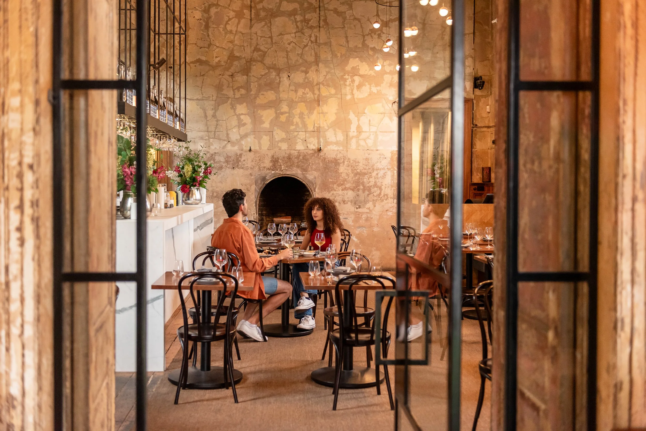 People dining in a cozy restaurant seen through a decorative iron gate and wooden frame, with exposed brick and textured wall behind them, and warm lighting overhead.