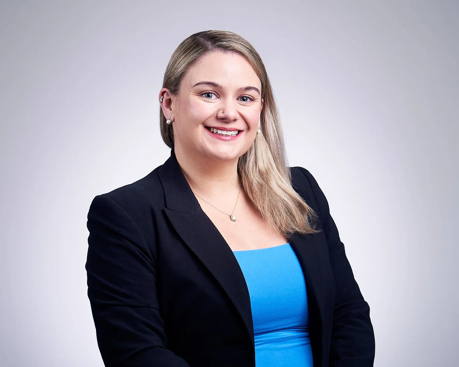 A woman with blonde hair smiling, wearing a black blazer and a blue top, against a plain gray background.