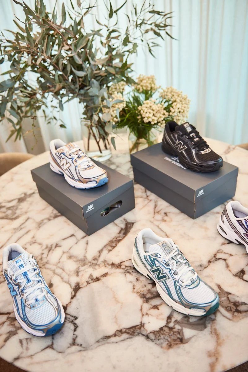 Collection of New Balance sneakers in black, white, and blue colors displayed on a marble table with boxes, with a plant and white flowers in the background.