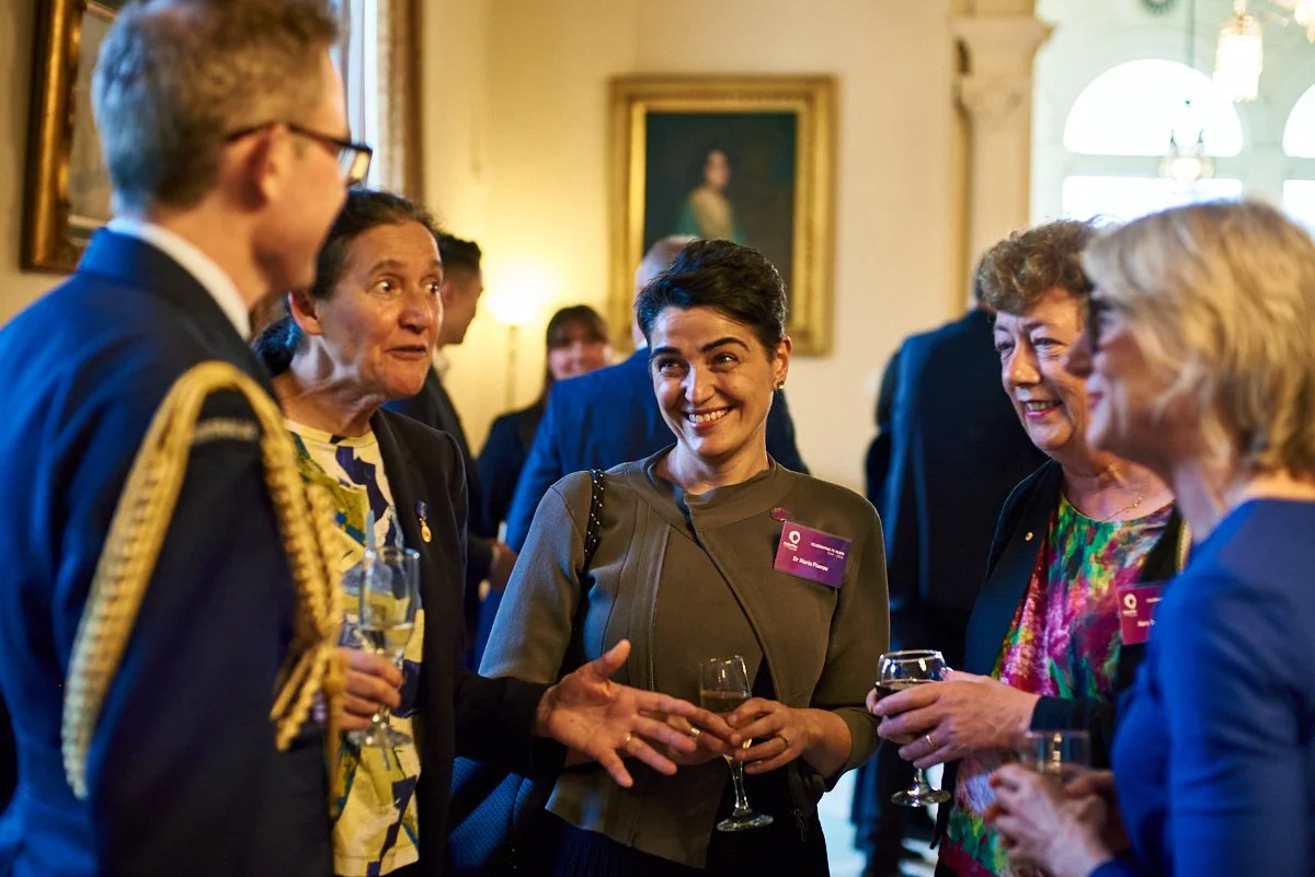 Group of professionally dressed women and a man engaging in conversation at a social event, holding wine glasses in an elegant room with artwork and large windows.