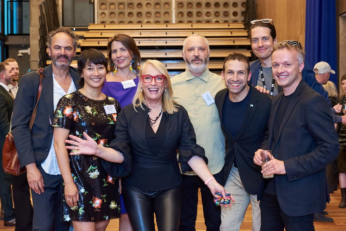 Group of nine diverse people smiling and posing for a photo at an indoor event, with wooden stairs and other attendees in the background.