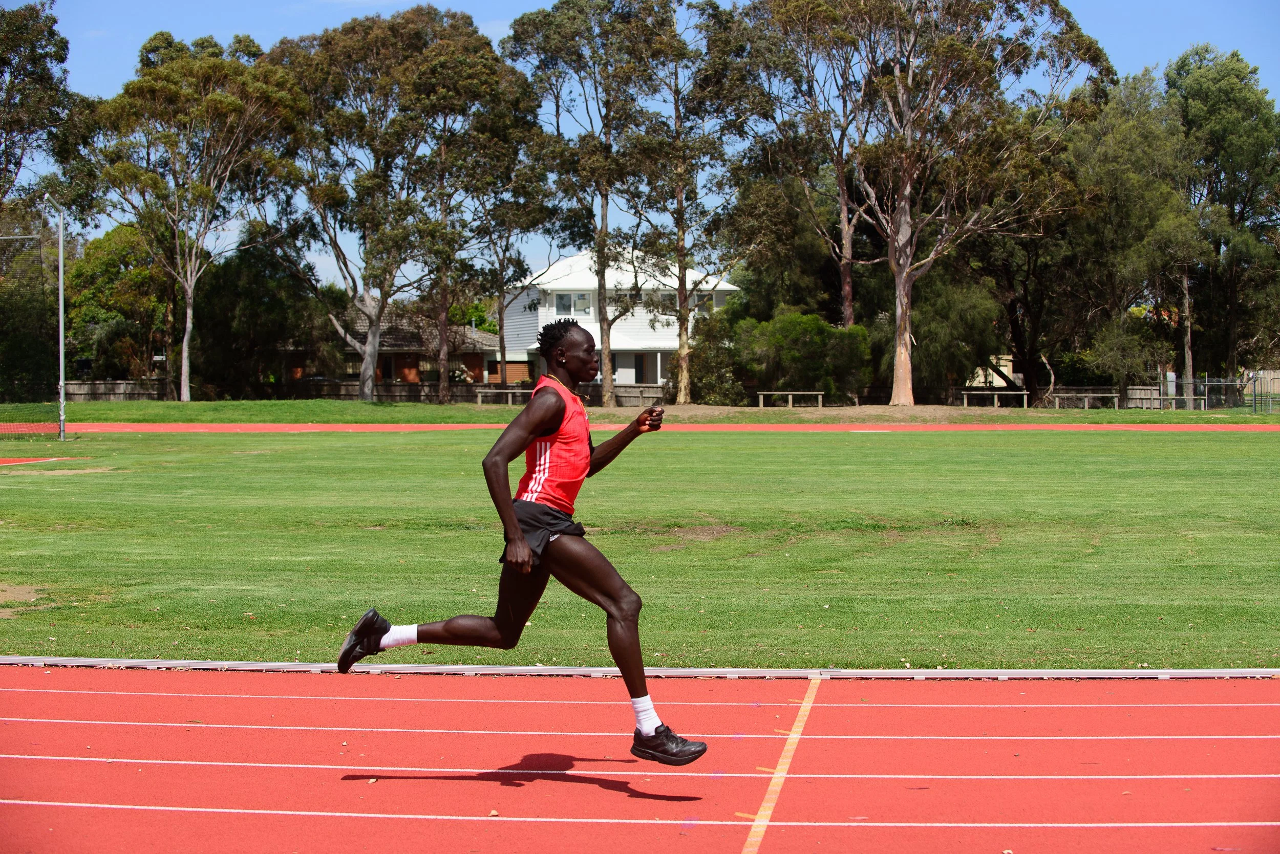 A male runner in a red tank top and black shorts running on a track field with trees and houses in the background.