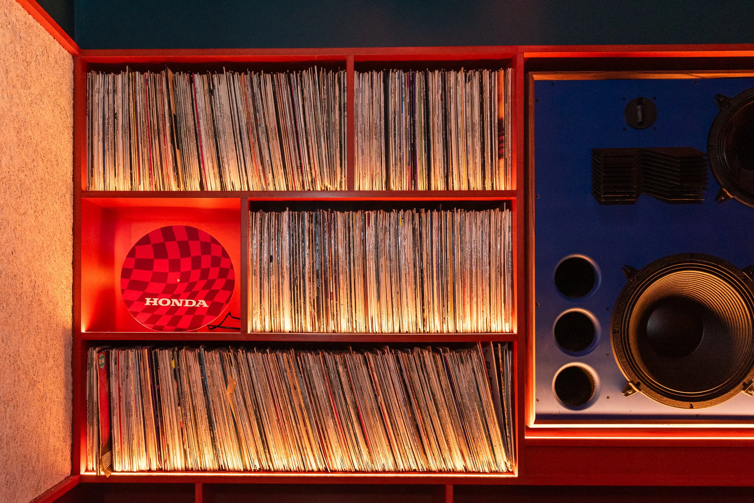 A red shelf filled with vinyl records, a red and black checkered Honda sign, and a large speaker in a room with dark walls and colorful circular wall decorations.
