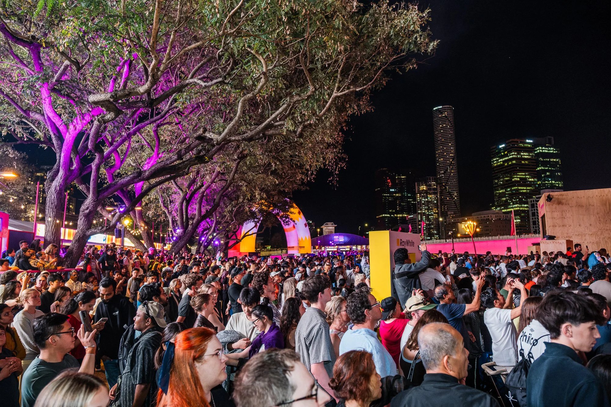 A large crowd at an outdoor event in an urban setting at night, with a prominent tree illuminated with purple lights on the left, and high-rise buildings with lit windows on the right. There are several tents and banners among the crowd.