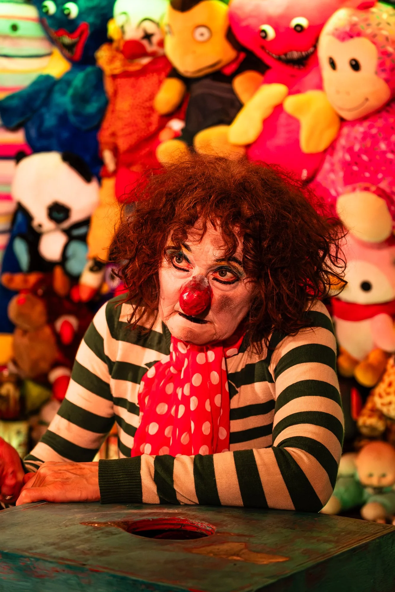 A person dressed as a clown with a red nose, striped shirt, and polka dot scarf, sitting at a table with a colorful painted hole in the surface, surrounded by colorful plush toys and stuffed animals in the background.