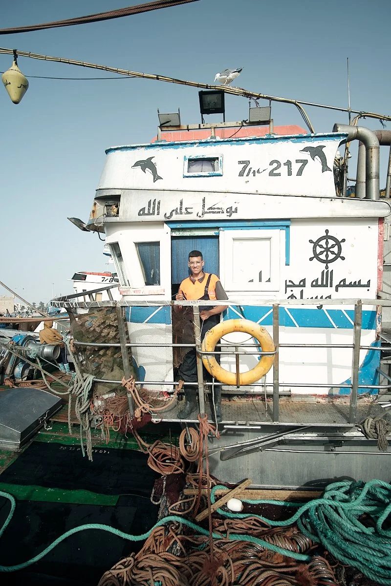 A young man standing on a boat docked at a harbor, with fishing ropes and equipment around him. The boat has writing in Arabic, dolphin illustrations, and a ship's wheel symbol on its white and blue exterior.